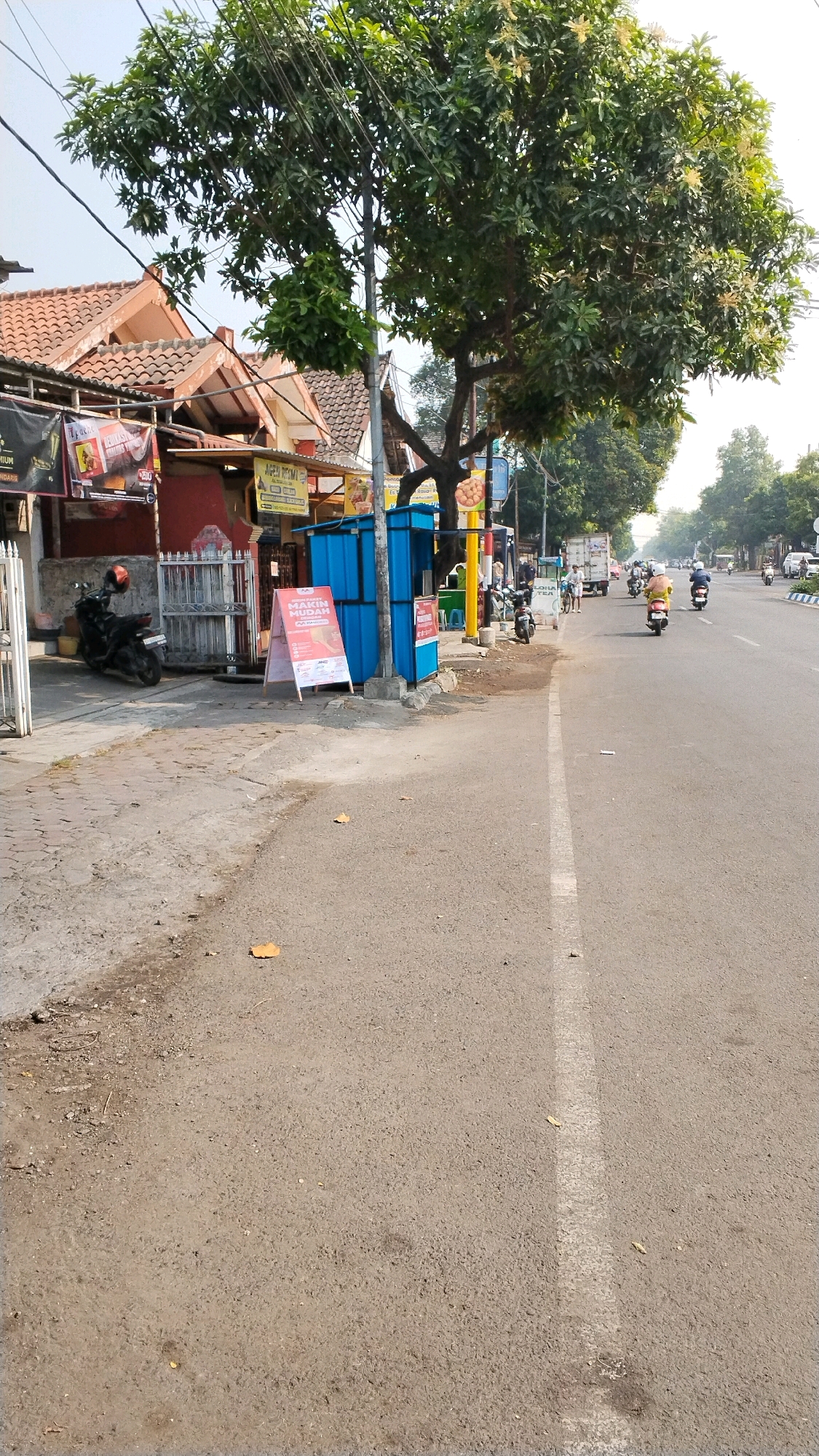 The image shows a street scene in Banjaragung, Indonesia. A large tree with dense green foliage dominates the left side of the frame, casting shadows on the adjacent buildings and sidewalk. The buildings appear to be commercial establishments with red and brown tiled roofs. A white picket fence separates the sidewalk from a small courtyard area where a black motorcycle is parked. Two blue utility boxes are positioned on the sidewalk, one of which has a red and white promotional sign in front of it. The street itself is paved and shows signs of wear, with a white line marking the lane. Several motorcycles, some with riders, are visible on the road, suggesting moderate traffic. The sky is clear and bright, indicating daytime.