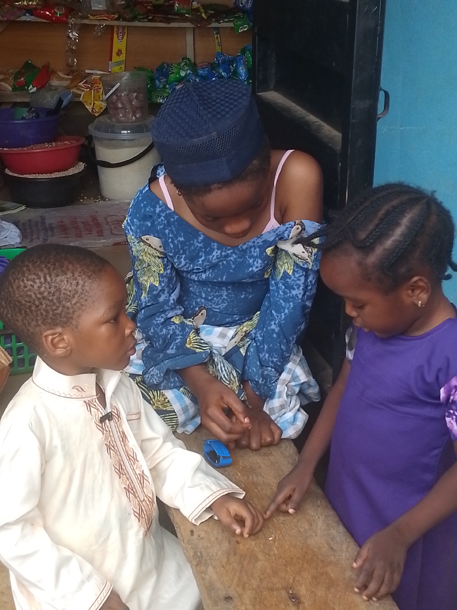 Three children are gathered around a wooden table in what appears to be an outdoor market stall in Abeere, Nigeria. On the left, a young boy wearing a white, embroidered traditional outfit sits facing right. In the center, a slightly older girl in a blue patterned dress and a navy blue headscarf looks down at the table, her hands resting on it. To the right, a young girl in a purple dress also looks down at the table, her finger pointing. The background shows shelves stocked with various goods, including bags of snacks, plastic containers, and buckets, suggesting a commercial setting. The overall scene depicts a moment of shared attention and interaction among the children in a busy market environment.