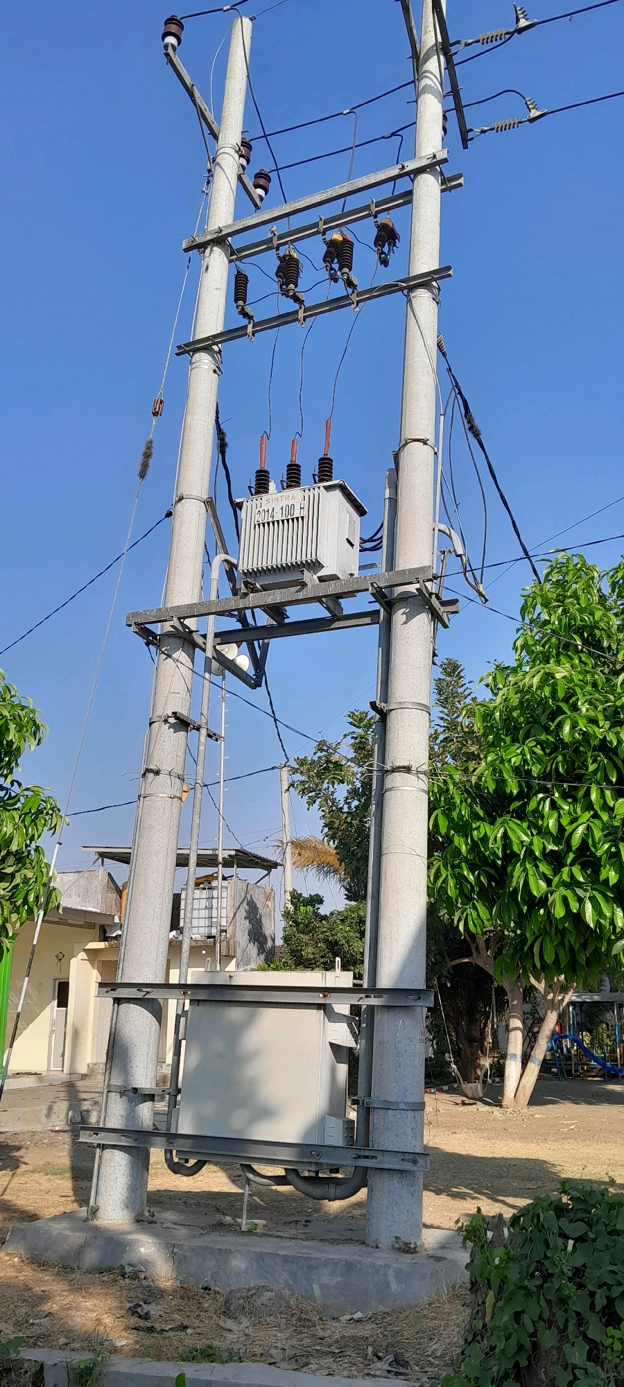 This image depicts an electrical transformer unit mounted on a tall pole structure in what appears to be an outdoor, semi-urban or rural setting, likely in Bendungan, Indonesia. The transformer, a grey, rectangular box with cooling fins and three prominent ceramic insulators on top, is attached to metal bracing supported by two substantial concrete poles. Wires are suspended from these poles, connected to various insulators and other electrical components.
In the background, there's a light-colored building with a pitched roof and a water tank visible on top, suggesting a residential or commercial area. Lush green trees frame the scene on the right and left sides. The ground is covered with dry grass, dirt, and some scattered debris. The sky above is a clear, bright blue. The overall impression is of everyday infrastructure in a tropical or subtropical climate. There are no people visible in the image.