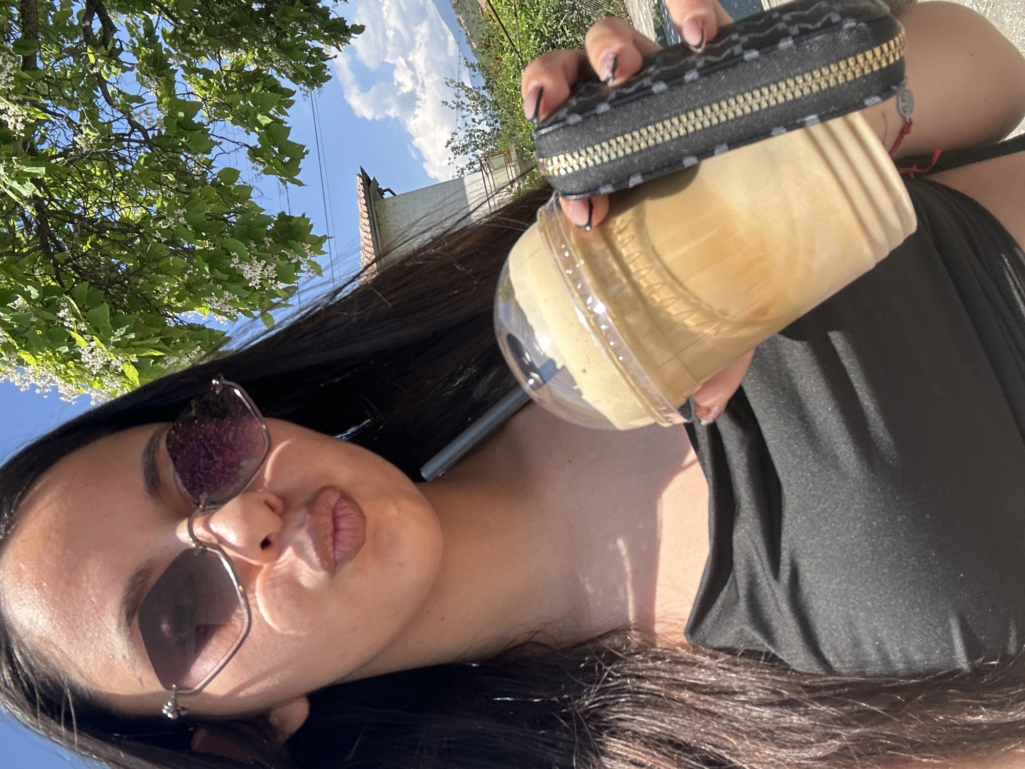 A selfie-style image, rotated ninety degrees counter-clockwise, features a person holding an iced coffee and a small zippered pouch outdoors under a sunny sky. The person, who has long dark hair and is wearing sunglasses, is looking towards the camera with puckered lips, making a kissing face. They are wearing a black top. Their hand, visible in the upper right, has manicured nails with dark tips, and a red bracelet or thread is around their wrist.
In the person's hand, a clear plastic cup filled with a light brown, frothy iced coffee drink is visible, topped with a dome lid. Above the coffee, a small black zippered pouch with a textured or patterned surface and a gold-colored zipper is held.
The background shows a vibrant blue sky with fluffy white clouds. A lush green tree with numerous small white blossoms fills the upper left portion of the frame, indicating a natural, possibly park-like or garden setting. Beyond the tree, parts of a building with a red-tiled roof and light-colored walls can be seen. Overhead wires are also faintly visible against the sky. The bright sunlight suggests it is daytime with clear weather. No visible text is present in the image.