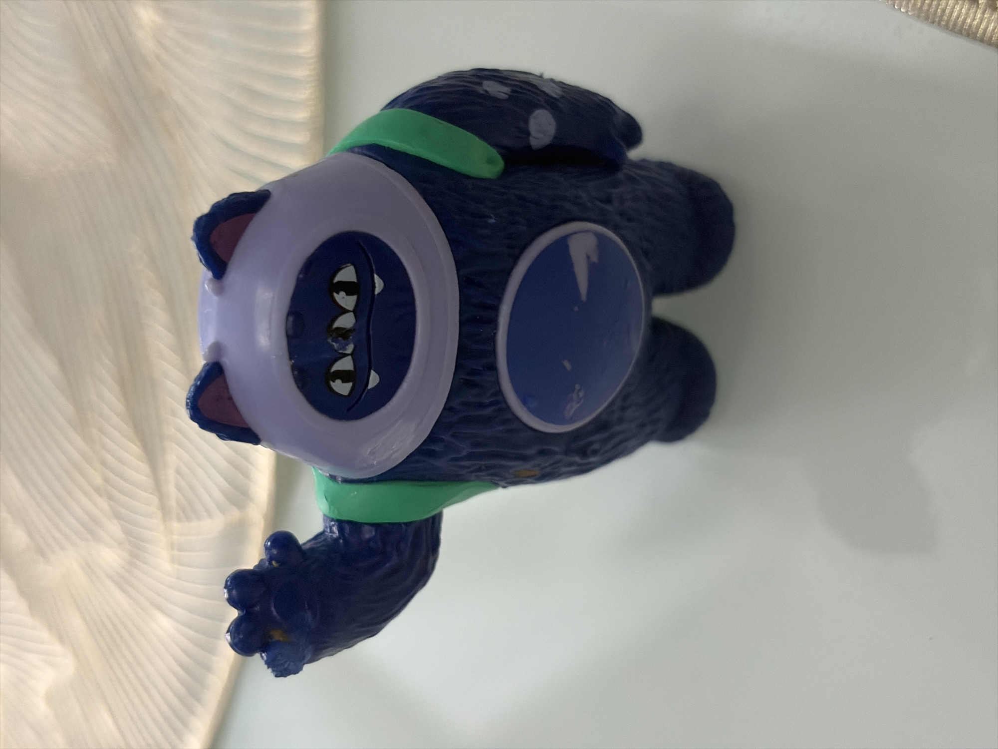 A close-up, slightly angled shot shows a blue, furry toy monster with a purple helmet-like head and green straps across its chest. The monster has three eyes, a wide mouth with visible teeth, and a single hand raised in a wave. The background is blurred and appears to be a light-colored, textured surface, possibly fabric. The monster is positioned on a smooth, white surface. The image is sharp and well-lit, with no discernible human presence or context suggesting a specific location in Moscow, Russia.