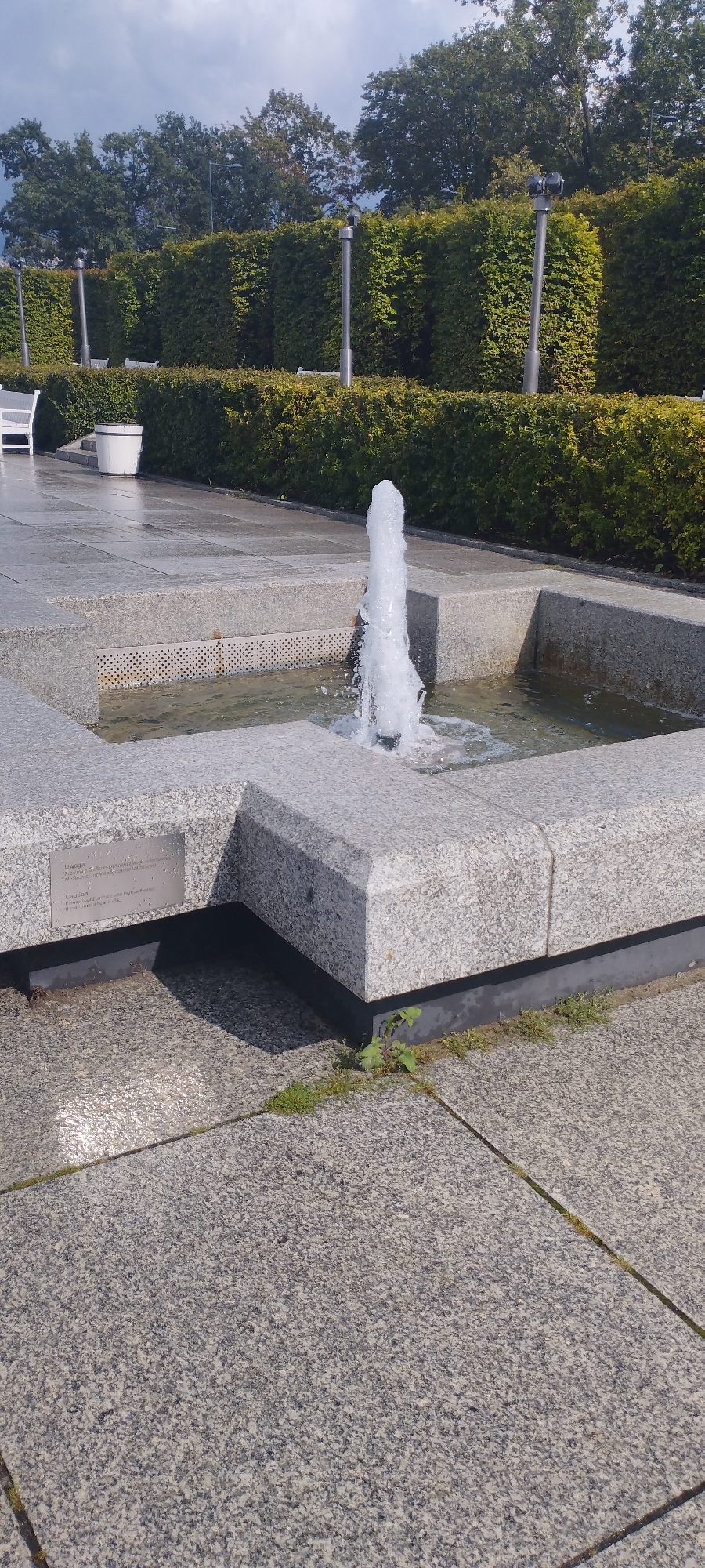 The image depicts an outdoor scene in Warsaw, Poland, featuring a modern fountain with a single water jet. The fountain is constructed from granite blocks and is surrounded by paved areas and manicured hedges. In the background, trees can be seen beyond the hedges. There are also white benches and a white cylindrical object, possibly a planter or waste bin, visible in the mid-ground. A small metal plaque is affixed to the side of the fountain, providing information or a warning, with text visible in both Polish and English. The lighting suggests it is daytime, with shadows cast on the pavement. The overall impression is of a well-maintained public space, likely a park or a plaza.