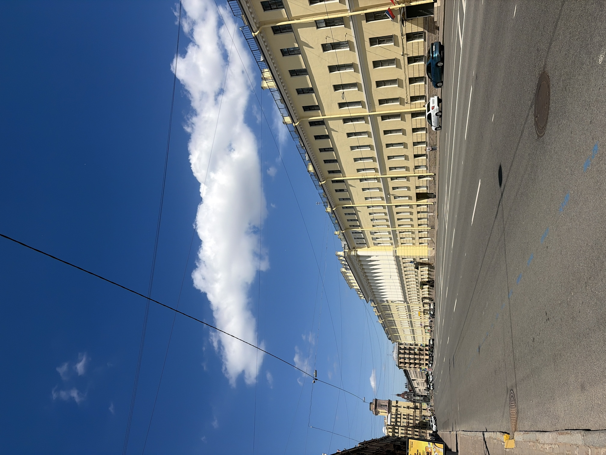 This image captures a daytime view of a wide, paved street in Saint Petersburg, Russia, with a prominent yellow building lining the right side. The building is multi-story, with a long facade punctuated by numerous windows and decorative architectural elements. A black metal railing runs along the roofline. Several cars are parked or driving on the street, including a white sedan and a teal sedan near the yellow building. A single person is visible standing near the yellow building. The street is marked with white lane lines and some blue dashes. The sky above is a vibrant blue with scattered white clouds, and power lines are visible stretching across the frame. On the left side of the street, other buildings of varying heights and architectural styles can be seen, along with a sidewalk and some advertising signs. The overall impression is an urban streetscape under a clear, bright sky.