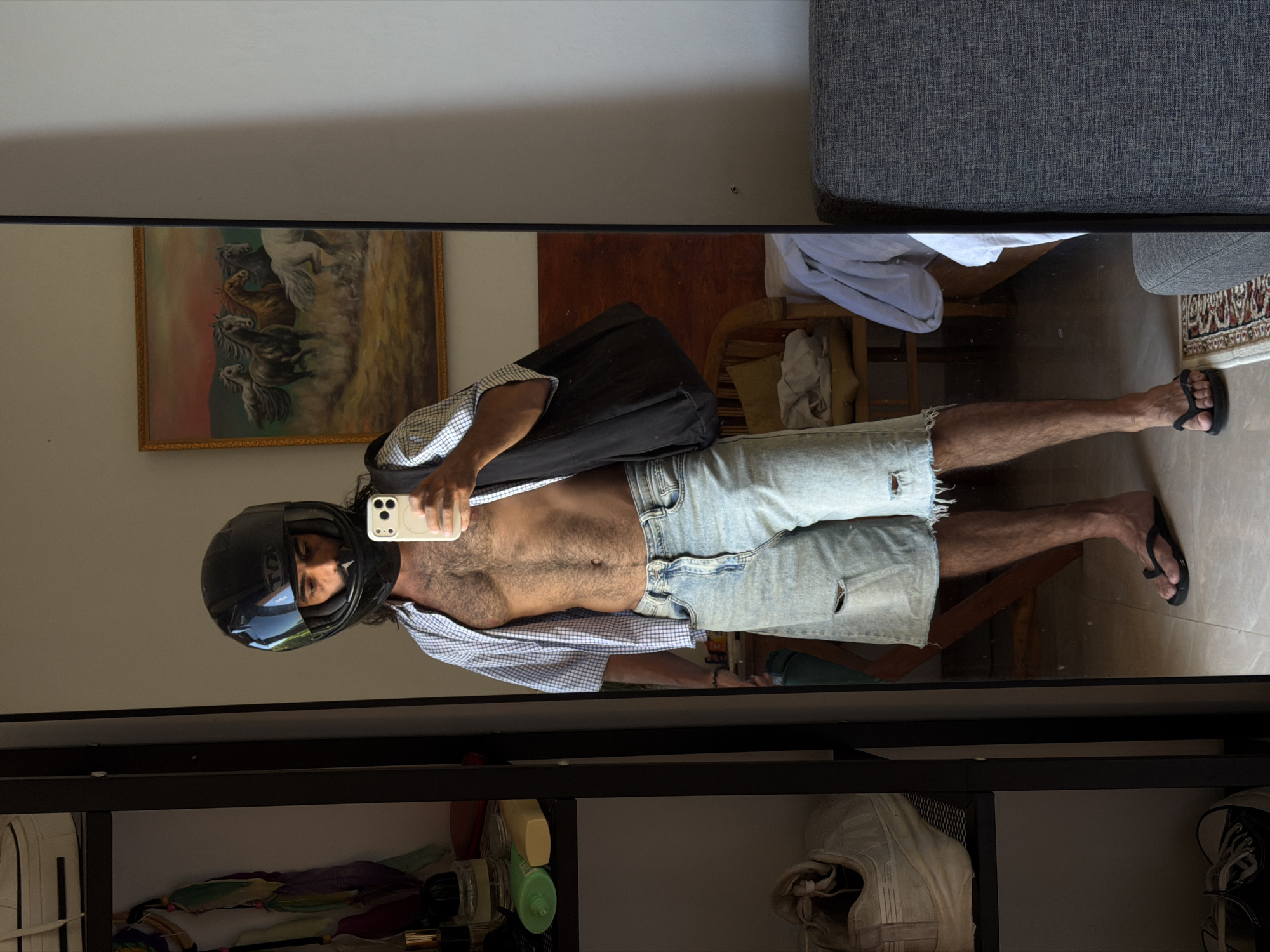 A person wearing a black motorcycle helmet, light blue denim shorts, and black flip-flops stands in front of a floor-to-ceiling mirror, holding a white smartphone. They are shirtless and wearing a partially unbuttoned checkered shirt, with a black bag slung over their shoulder. The background reflects a room interior with a wooden chair, a framed painting of horses, a gray sofa, and a patterned rug. A black shelving unit is visible at the bottom of the frame, containing various items. The location is North Kuta, Indonesia.