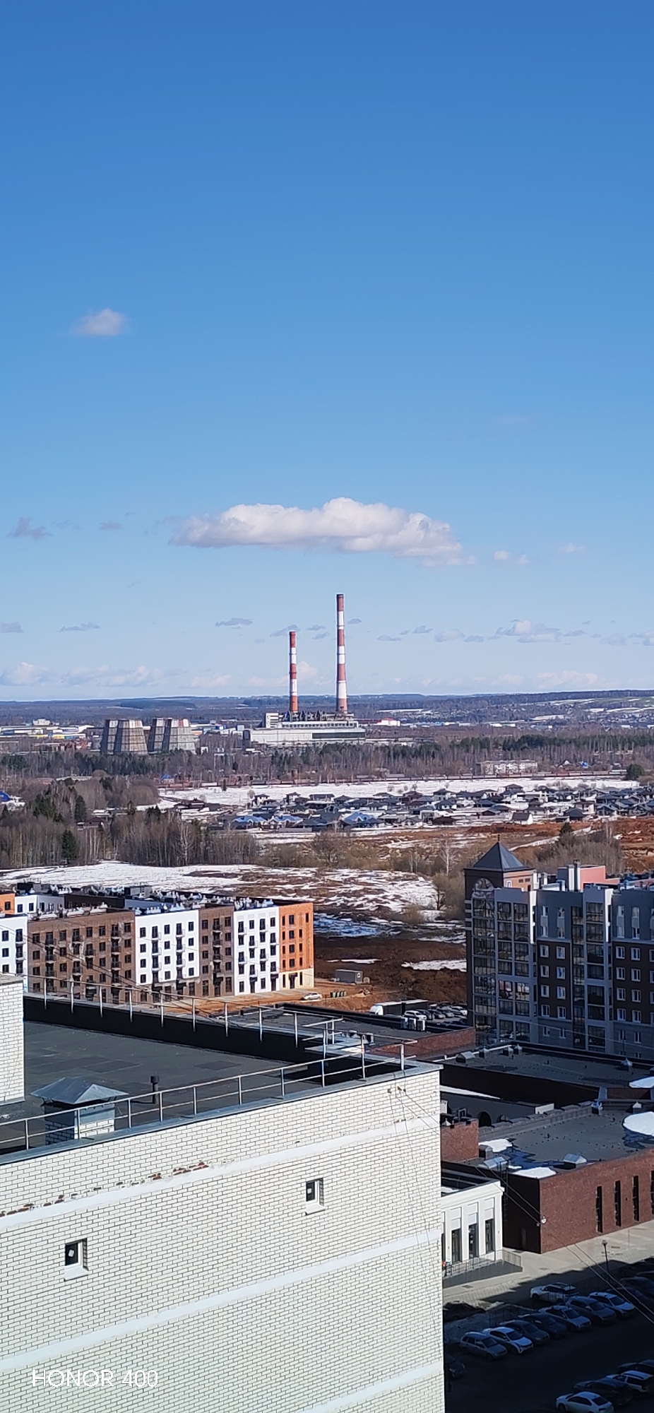 The image presents a high-angle view of a section of Izhevsk, Russia, on a clear day with a blue sky sparsely dotted with small clouds. The foreground is dominated by rooftops of buildings constructed of light-colored brick. Mid-ground features several multi-story apartment buildings, some with distinctive red brick facades. The ground between the buildings has patches of melting snow. In the background is a power plant identified by its cooling towers and two tall chimneys painted with red and white stripes. The landscape surrounding the power plant is a mix of forest and smaller buildings, characteristic of a suburban or industrial area.