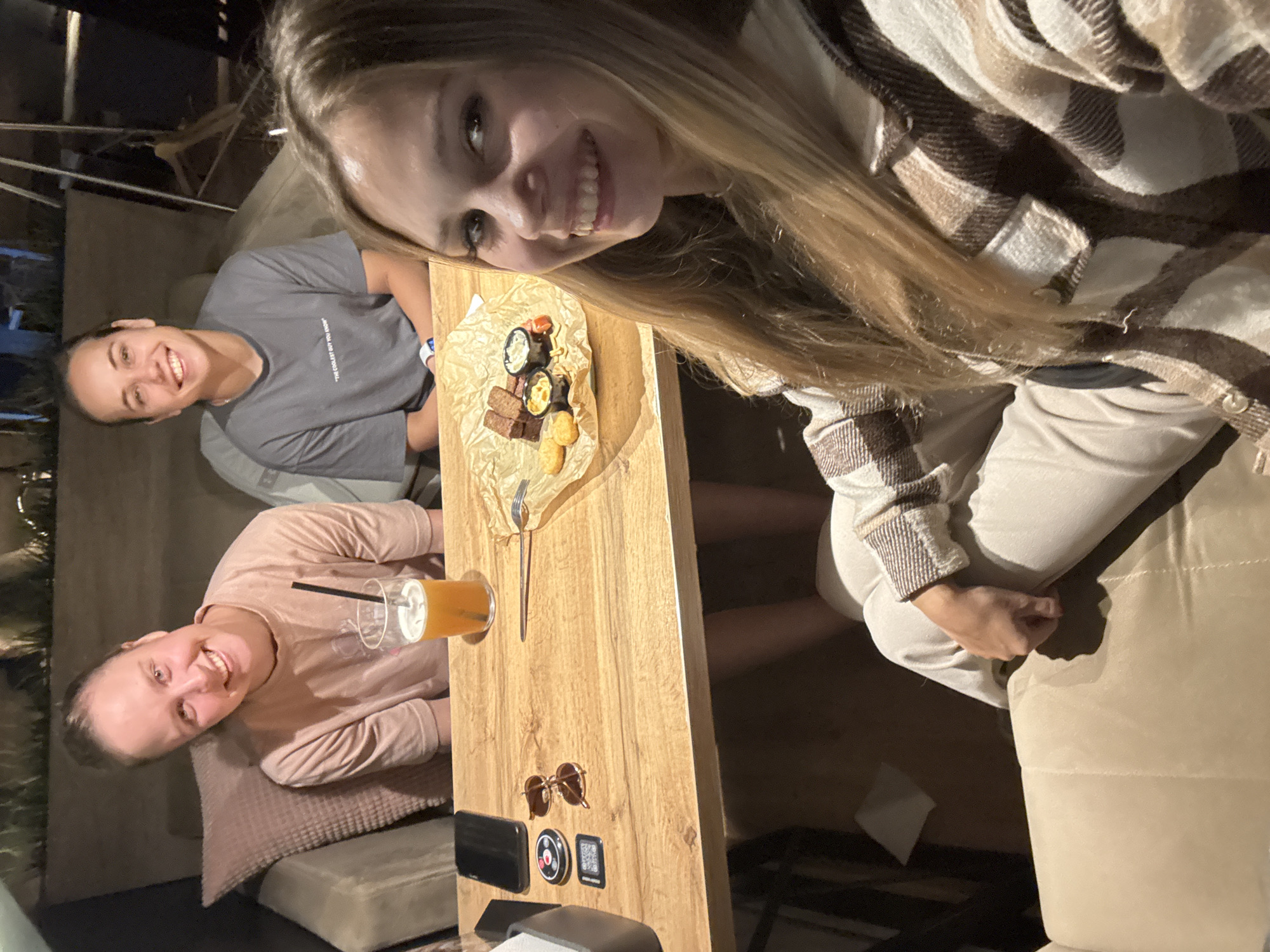 The image captures a candid moment with three young women gathered around a wooden table. Two of the women are seated, while the third is positioned at the top of the frame, leaning in towards the camera and smiling broadly. The setting appears to be an outdoor or semi-outdoor space, possibly a cafe or restaurant, with subdued lighting suggesting it is evening or nighttime.
On the table, there's a small plate of appetizers, including what looks like meat cubes, crackers, and dips, presented on brown parchment paper. A fork rests on the paper, ready for use. A clear plastic cup containing an orange beverage and a straw is also on the table, held by one of the seated women.
The subjects are dressed casually. The woman at the top is wearing a plaid shirt over a lighter-colored top. One of the seated women is in a grey t-shirt with visible text that reads "THE COOLEST GUY YOU KNOW". The other seated woman is wearing a pink long-sleeved shirt.
Other objects on the table include a pair of round, brown-tinted sunglasses, a black rectangular object (possibly a phone or power bank), and two small square items. The overall mood is relaxed and joyful, with all three women displaying pleasant expressions. The presence of drinks and snacks suggests a social gathering or meal. The location context of Chaikovsky, Russia, provides a geographical anchor, though there are no specific visual cues in the image to confirm this location.