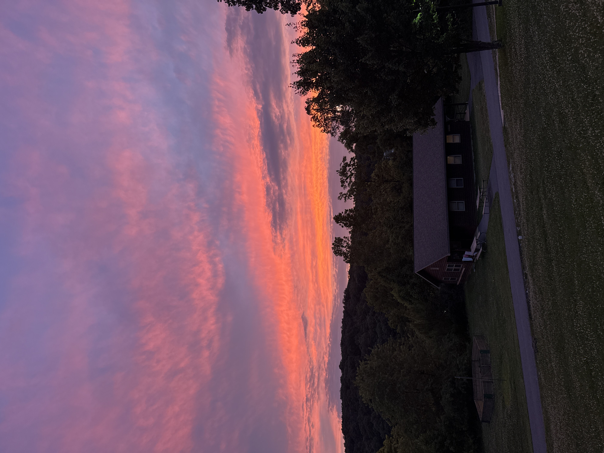 A wide-angle, eye-level shot captures a vibrant sunset over a rural landscape. The sky is a dramatic canvas of pink, orange, and purple hues, with wispy clouds stretching across the horizon. Below the sky, a dense line of dark green trees forms a silhouette against the colorful sky, with rolling hills visible in the distance. In the foreground, a brown, single-story building with a dark roof sits on a grassy area. A paved path leads towards the building, and a picnic table is visible in front of it. To the left of the building, a hexagonal wooden structure is present. The ground is covered in green grass dotted with small white flowers. A tall tree with dark green foliage stands to the right of the building, partially obscuring a streetlight. The scene appears to be a park or campground setting in Washington Township, USA.
