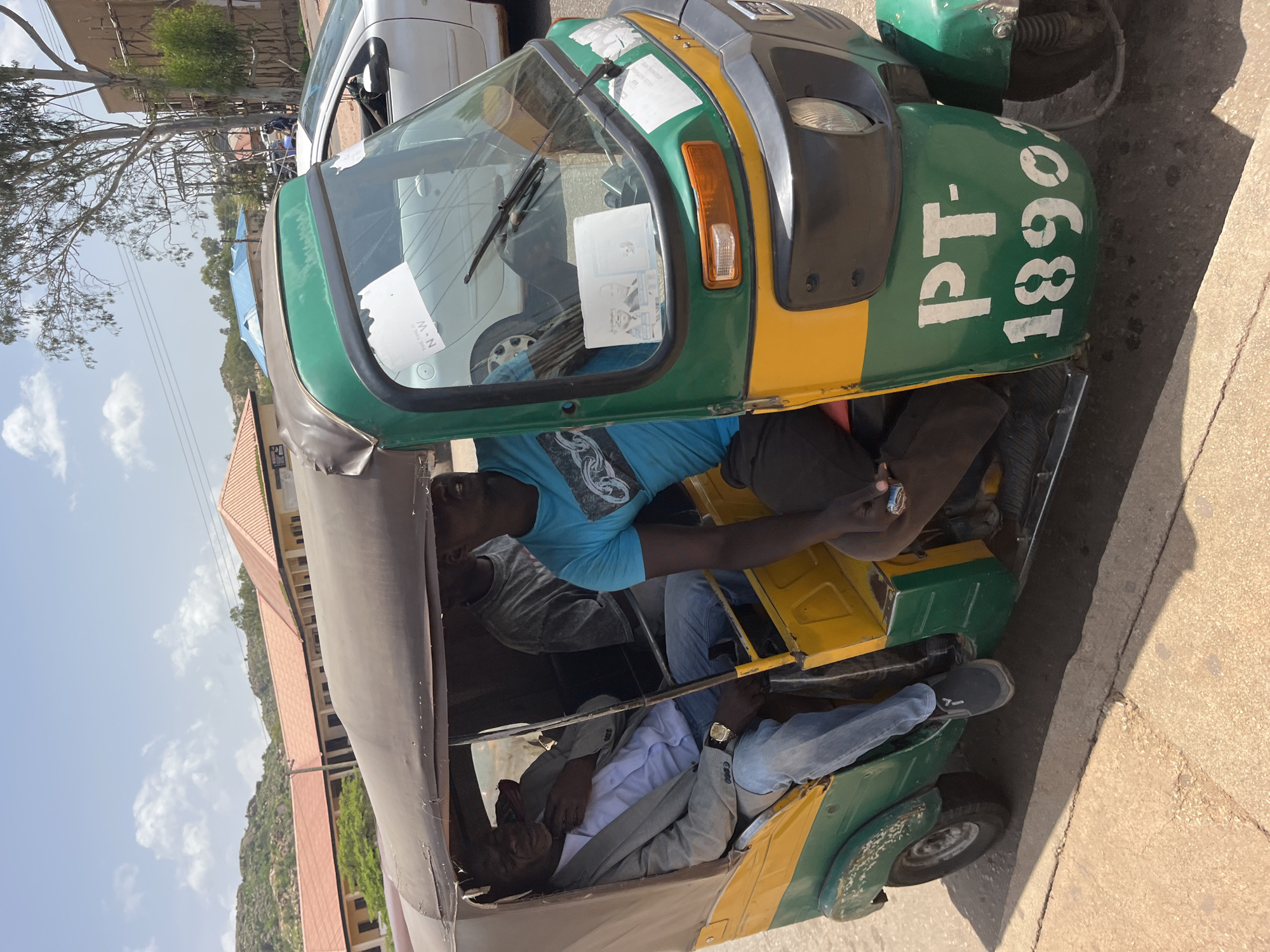 This media file shows a tuk-tuk parked on a paved surface, with a building and trees in the background. Three people are visible inside the tuk-tuk. One person, wearing a blue t-shirt, is in the driver's seat. Another person, wearing a light grey jacket over a white shirt, is seated in the passenger area. A third person, wearing a grey t-shirt and jeans, is also in the passenger area. The tuk-tuk is green and yellow, with visible white text on the back that reads "PT-1890". The scene appears to be outdoors during daylight, with a bright sky and some clouds. The overall atmosphere suggests a typical day in a populated area.