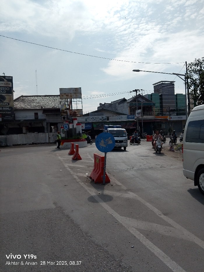 The image shows a street intersection in Jatibarang, Indonesia, at the coordinates -6.466 latitude and 108.3 longitude. There are several buildings on either side of the street, with the nearest being a white building with a red roof. The buildings appear to be mostly residential. The street is paved with asphalt and has white lane markings. There is a white van parked at the far right side of the image, partially obscured by the right edge of the image. A person is walking on the street. A blue and white traffic sign is centered in the image on a post. A white van is stopped in the street, with its front end facing right. On the far left, there is a red-and-white construction barrier. There is a small building, possibly a kiosk, with a sign that reads "BANK MEGA". Another building is visible in the background, with a sign that reads "WARTE". The sky is partly cloudy, with the sun shining brightly. The ground is dry. There is a person on a motorcycle in the center of the image. Two other motorcycles are parked on the street. A person in a red shirt is walking near the curb on the far left side of the image.