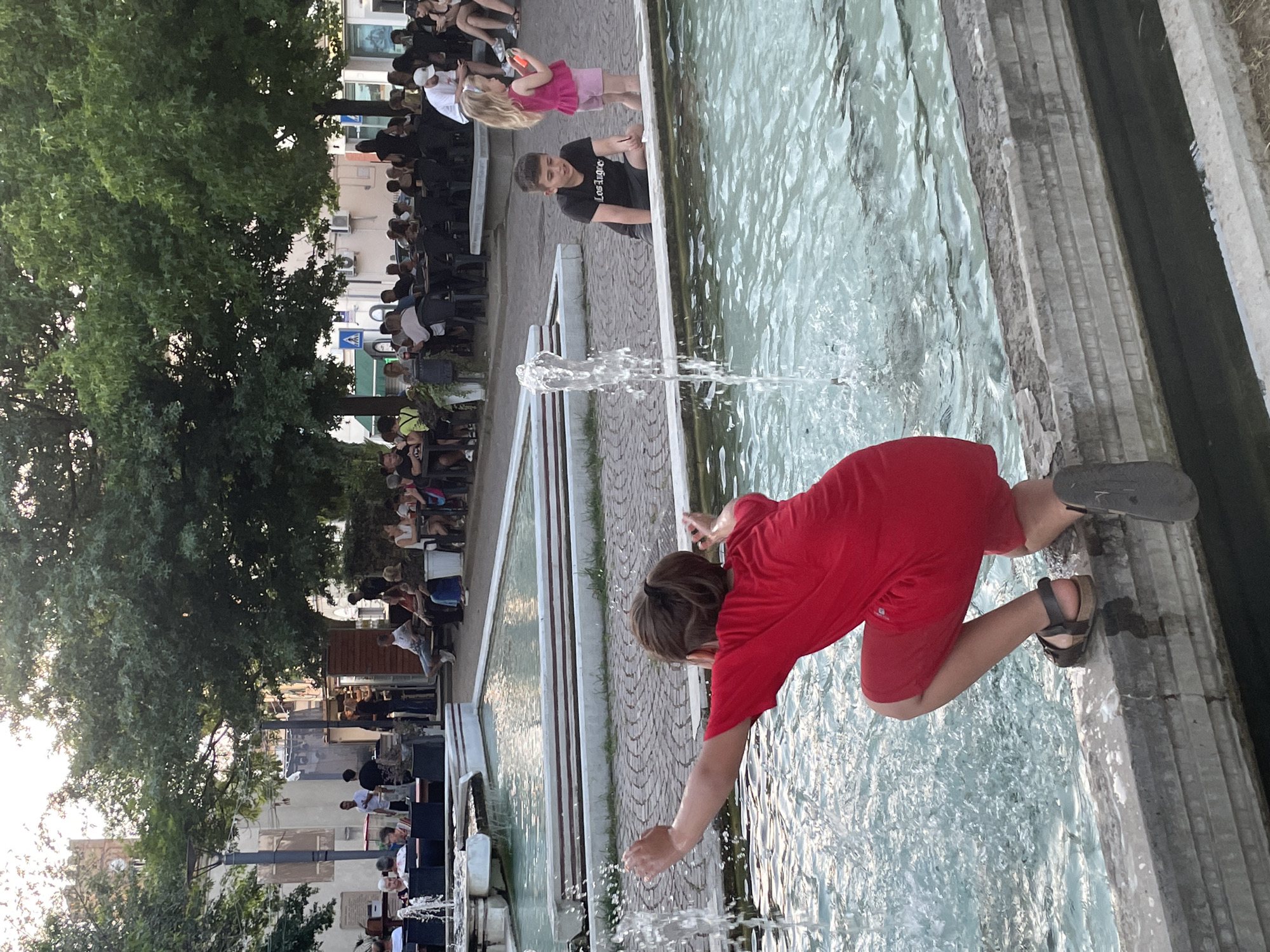 In Altopascio, Italy, a young boy in a red shirt and shorts is crouched at the edge of a fountain, splashing water with his hands. To his right, a boy in a black t-shirt with "Los Angeles" written on it and a girl in a pink top and shorts are standing near the fountain. Behind them, people are seated at tables outside, and trees provide shade. The scene appears to be a public square with a fountain, suggesting a social gathering or a recreational area.