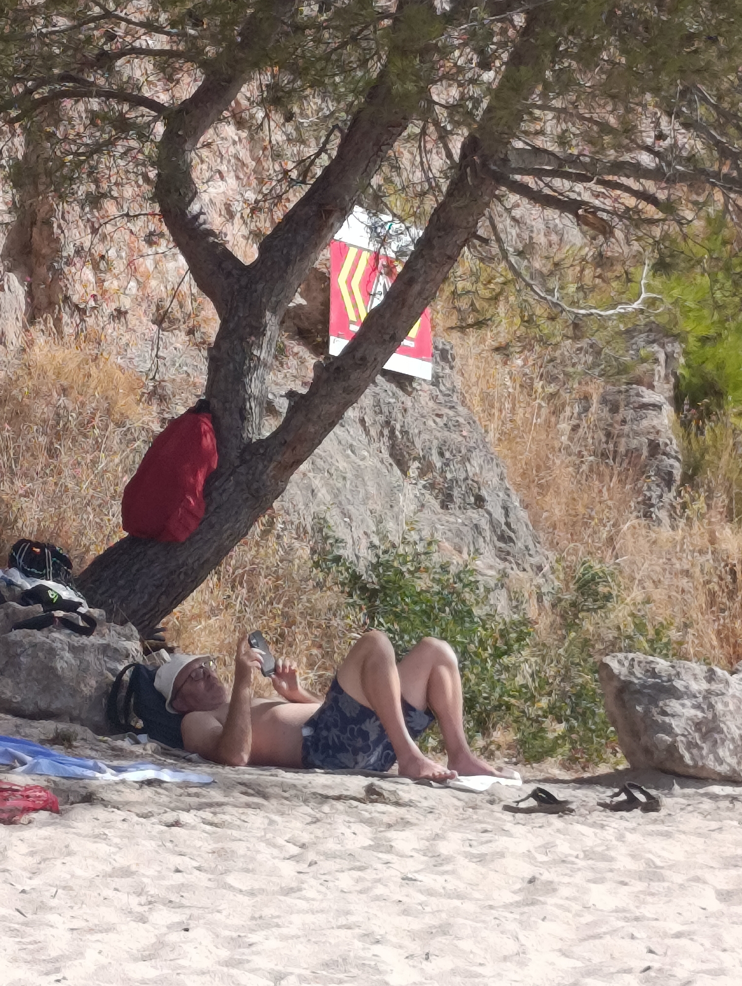 A man, wearing a white cap, sunglasses, and patterned swim trunks, reclines on a sandy beach in Setúbal, Portugal. He is lying on a towel, propped up on his elbows, and appears to be looking at his phone. His legs are extended in front of him, and a pair of dark sandals lie on the sand to his right. Beside him, to his left, are more items of clothing and a backpack, one resting on a rock and another leaning against a tree. A bright red backpack is also visible, slung over a branch of the tree. The tree itself, with a rough bark texture, casts a shadow over the man and the beach. Behind the tree, a rocky cliff face rises, sparsely covered with dry grasses and some green foliage. A red and yellow warning sign is attached to the tree. The scene suggests a casual, outdoor recreational setting on a sunny day.
