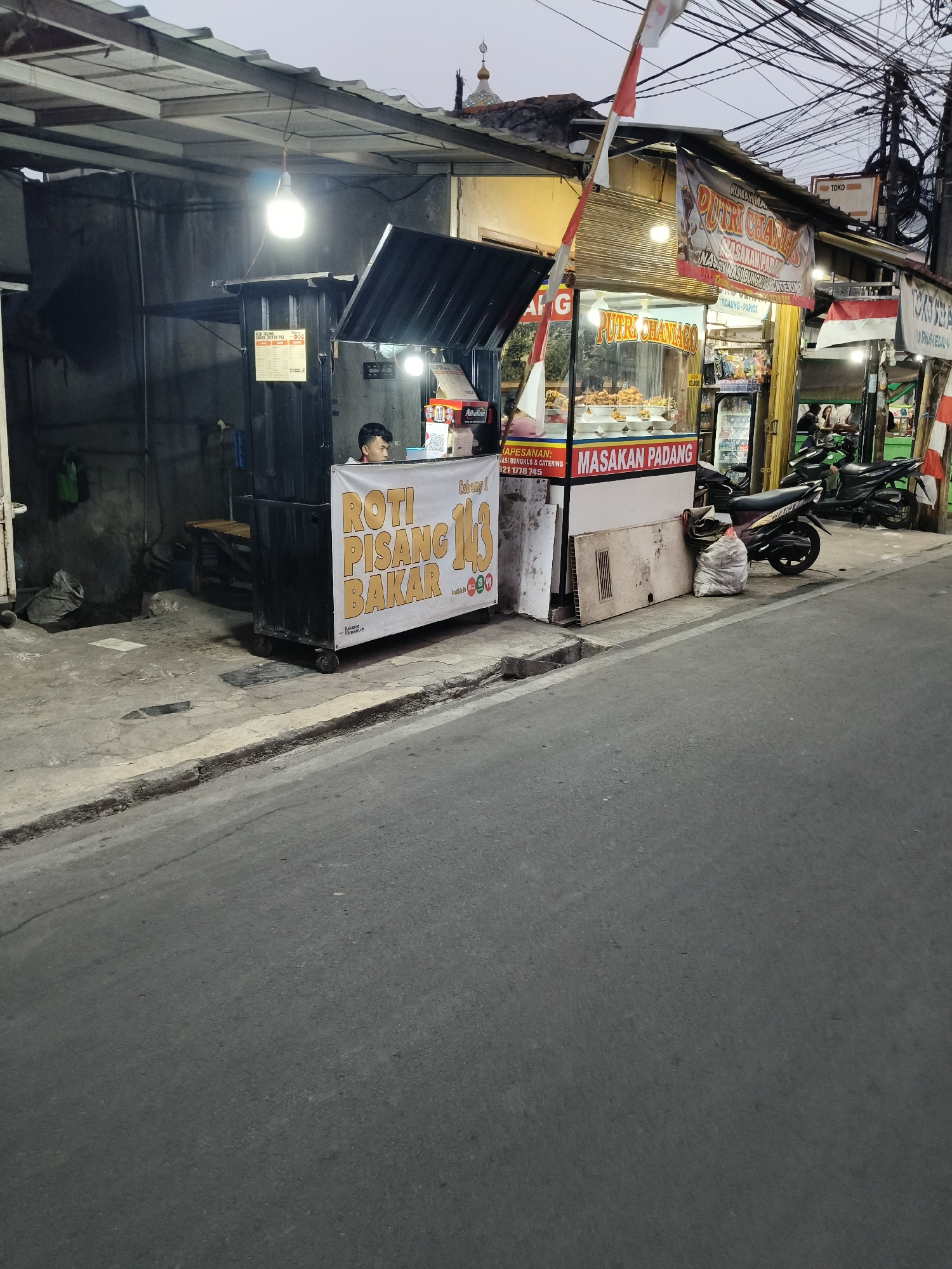 The image depicts a street-level view of a food stall in South Tangerang, Indonesia. The stall, identified by signage as "PUTRI CHANAGO" and "MASAKAN PADANG," appears to sell Indonesian cuisine, specifically Padang food. In the foreground, the asphalt road is visible, with a concrete curb and sidewalk. The stall itself is constructed from a dark container-like structure with an awning and a display area showcasing food items. A young man is seen serving customers or preparing food behind the counter. A prominent banner in front of the stall reads "ROTI PISANG BAKAR," indicating another item offered. Several motorcycles are parked nearby, and electrical wires hang overhead, typical of urban Indonesian streets. The overall scene suggests a bustling local eatery in an urban environment.