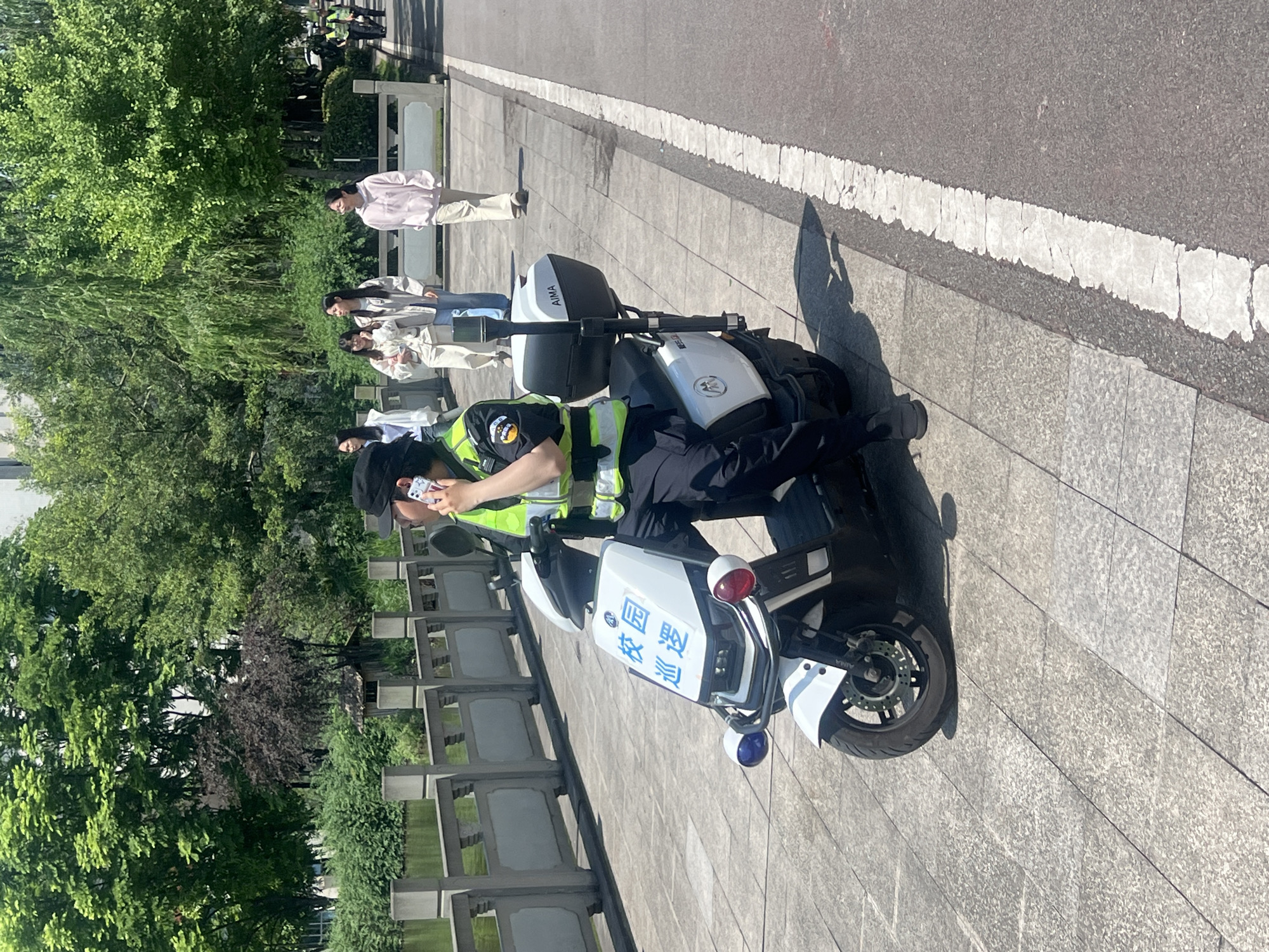 A police officer in a black uniform and fluorescent green vest sits on a white electric scooter. The scooter has Chinese characters that read "Campus Police Station" and a blue light below. The officer is holding a phone to their ear. A group of four people are standing in the background near trees and a low stone wall. The scene appears to be outdoors on a paved area next to a road with a white line. The location is Jiangjin, China.