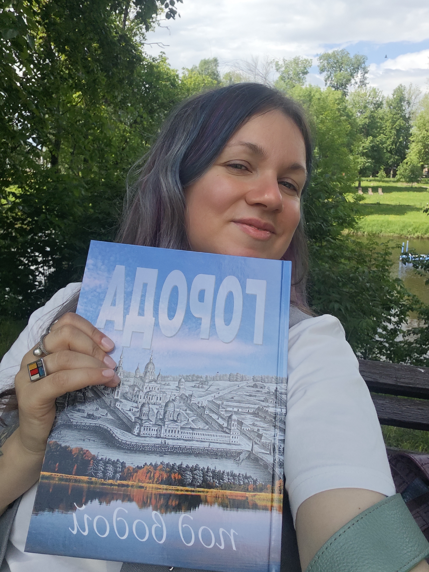 A woman with light skin and dyed hair with streaks of blue and purple is holding a book in front of her chest while seated outdoors. The book has a light blue cover with a drawing of a historical building complex and a lake. The text on the cover reads "ДОБРОСЕЛЬЕ" ("Dobroselye") in large white letters, and "Слободской бол" at the bottom. The woman is wearing a white shirt with a light gray vest. She is wearing a ring with a colorful square design on her left hand. She is sitting on a bench in front of a grassy area, with lush green trees in the background. In the distance, a pond or river can be seen. The photo was likely taken in Selivanovo, Russia, where the Dobroselye church is located.