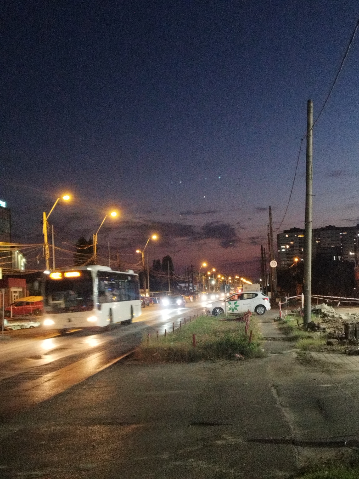 The image depicts a street scene in Bucharest, Romania, at dusk. A white bus, possibly a public transport vehicle, is in motion on the left side of the road, its headlights illuminated. Several streetlights are visible, casting a warm glow on the wet asphalt. A white car with a green logo is parked on the right side of the road, and a speed limit sign indicating "30" is posted nearby. In the background, apartment buildings with illuminated windows suggest a residential area. The sky is a deep blue transitioning to hues of purple and orange near the horizon, with some clouds present. The road appears to be undergoing construction or maintenance, with dirt piles and barriers visible on the right side. The overall impression is of an urban environment at the end of the day.