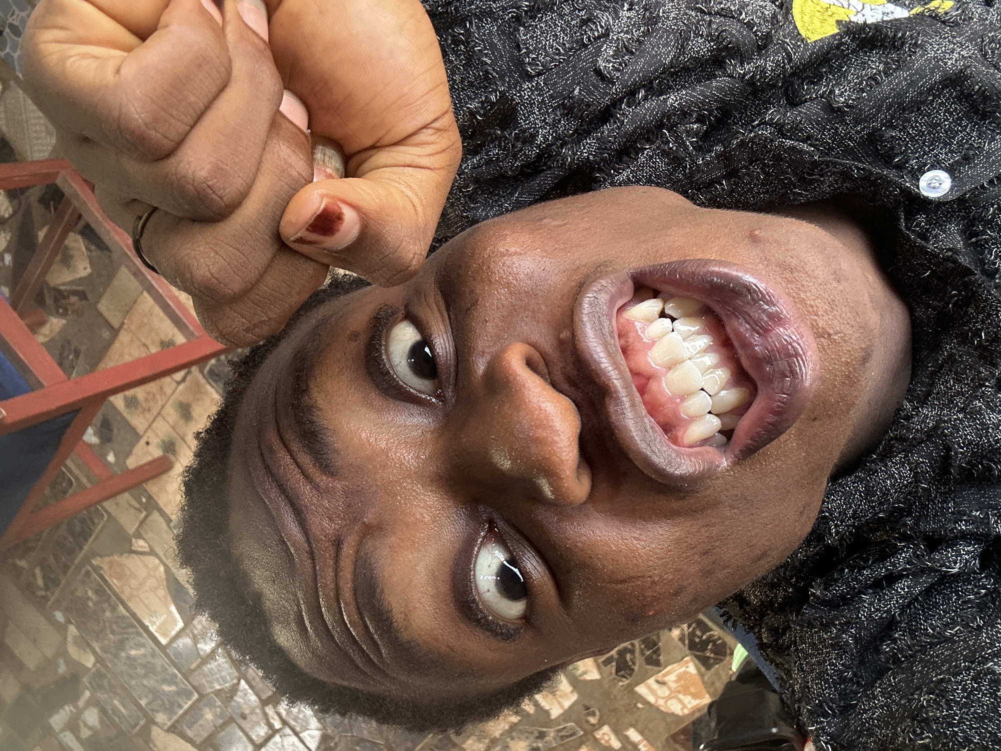 A close-up, low-angle shot focuses on a dark-skinned individual with their head tilted back, showing their open mouth and teeth. The person has a serious expression with both eyes visible. They are wearing a dark-colored woven shirt or sweater with a button visible near the collar. The individual's left hand is positioned near the top left corner of the frame, with a ring on the finger and a nail with dark nail polish. The background consists of a tiled floor with a geometric pattern and a portion of a red-painted chair or stool. The location is identified as Unknown, Unknown.