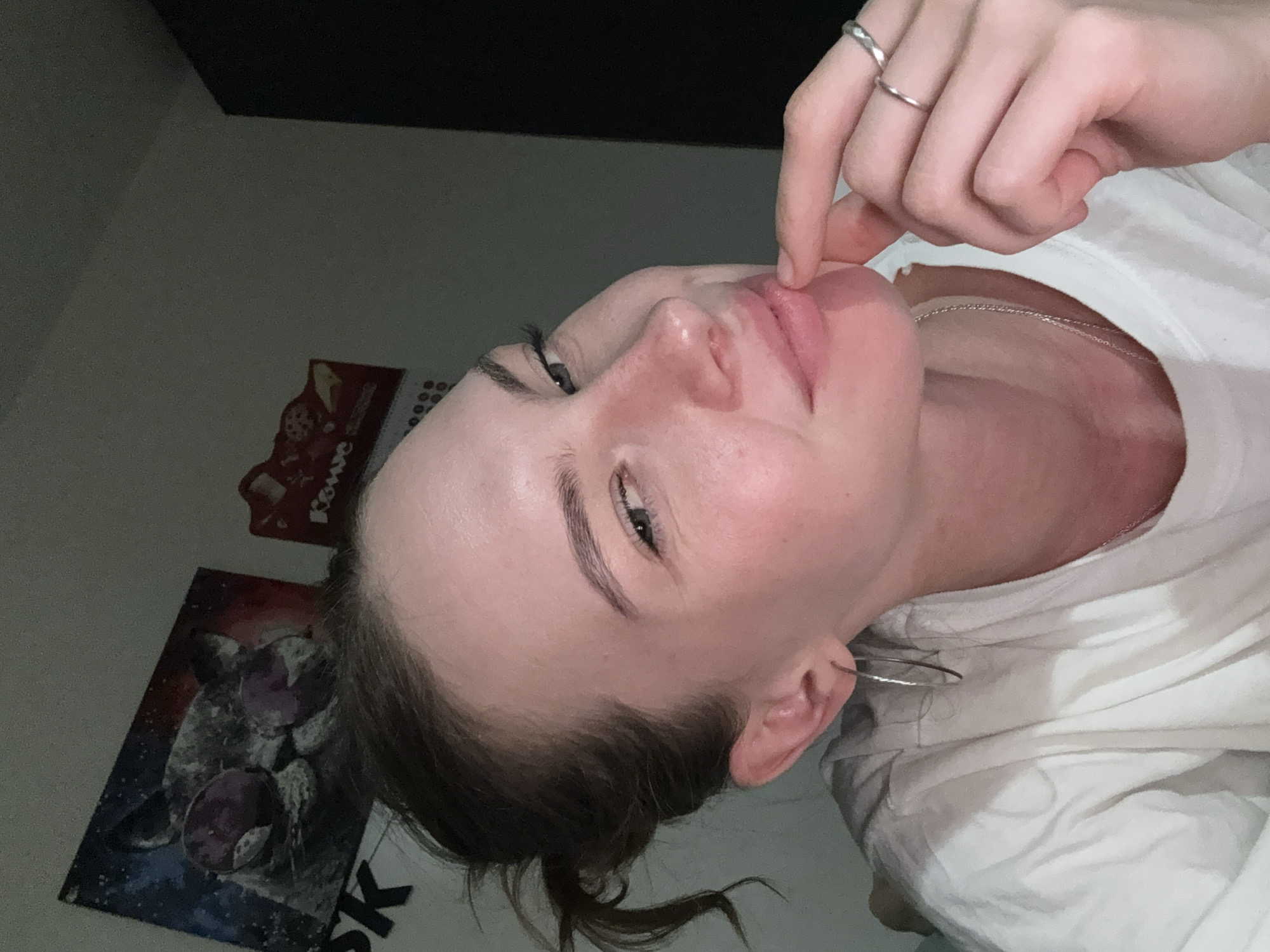 In a dimly lit room, a young woman with light skin and brown hair tied back in a bun is captured in a close-up selfie. She is wearing a white t-shirt and several pieces of jewelry, including hoop earrings, a silver necklace, and two rings on her right hand. Her left hand is resting on her chin, while her right hand is near her lips. The background reveals a wall with two posters: one of a cat wearing sunglasses and another with what appears to be a calendar or decorative item. The lighting suggests it is evening or night. The image is a selfie, taken by the woman herself. No specific location in Perm, Russia, can be determined from the visual information provided.