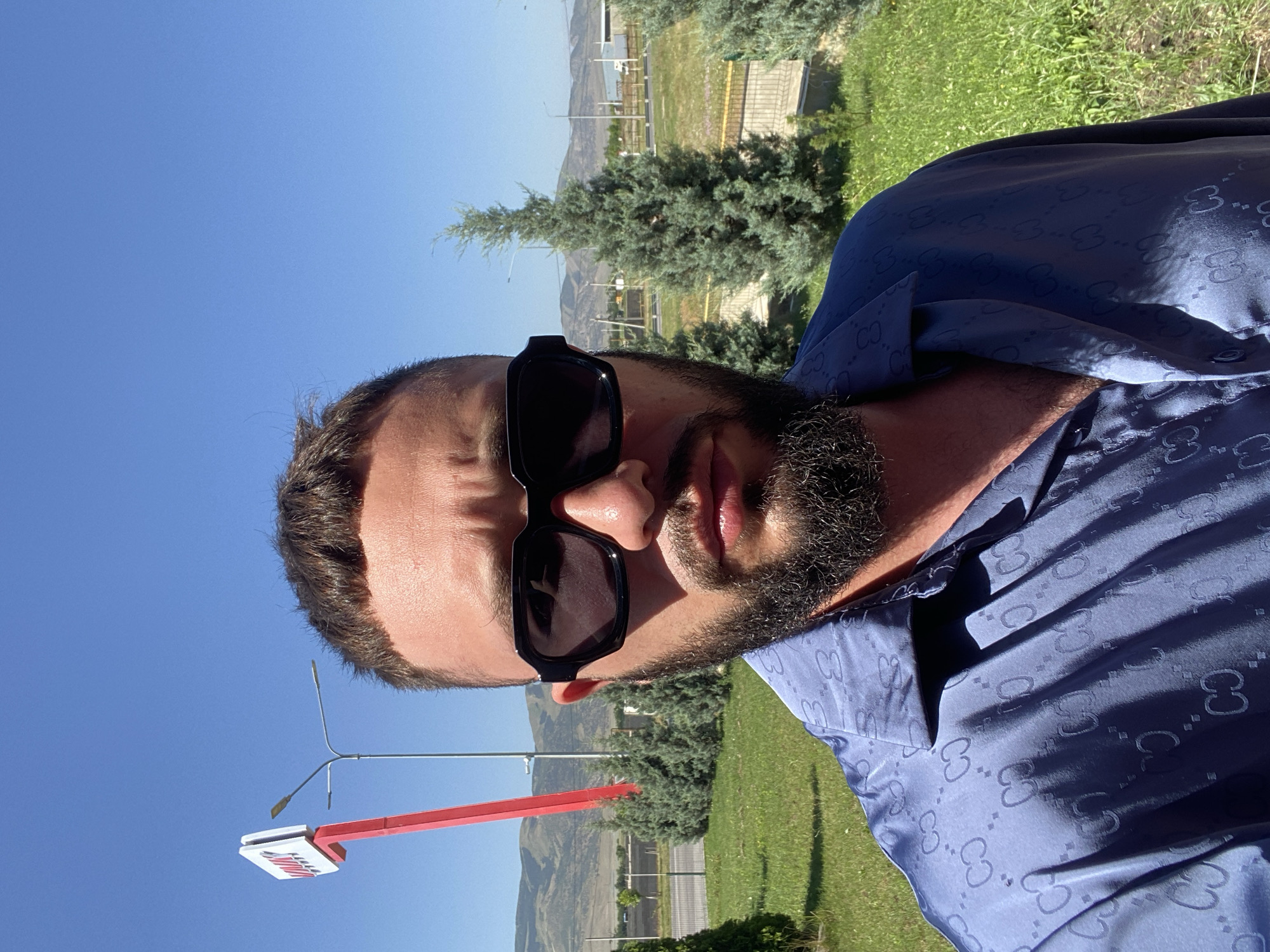 A man with dark hair, a beard, and sunglasses is taking a selfie outdoors in the city of Korkmaskala, Russia. He is wearing a blue patterned shirt. Behind him, there are trees, a red sign with white lettering on a pole, streetlights, a road, and hills in the distance. The sky is clear and blue, and the sun is shining.