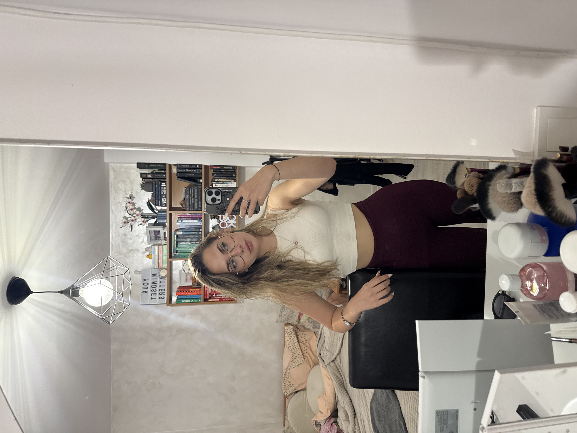 A woman is taking a mirror selfie in a room. She is wearing glasses, a white cropped top, and maroon leggings. She is holding a phone with a patterned case in her left hand. The room appears to be a bedroom, with a bed covered in blankets and pillows visible in the background. There is a bookshelf filled with books behind her, and a decorative lamp is hanging from the ceiling. The image also shows a vanity table with makeup brushes and various cosmetic products. There is no visible information to identify if this is in Blagoevgrad, Bulgaria.