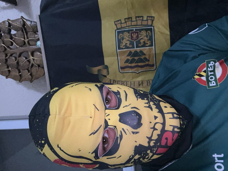 The image depicts a single individual in a frontal close-up, situated indoors in the city of Sedlartsi, Bulgaria. The person is wearing a yellow and black balaclava featuring a stylized skull design, with eyeholes revealing their brown eyes. Red text, partially legible as "12TR", is present on the lower portion of the mask. The individual is clad in a dark green long-sleeved shirt adorned with a circular logo on the right chest, identified as that of FC Botev Plovdiv, a Bulgarian football club, featuring "БОТЕВ 1912". Behind the individual, a black and yellow flag is draped, displaying the coat of arms of Plovdiv, Bulgaria, and partially visible Cyrillic text "ДРЕВЕН И В", referencing Plovdiv. Above the flag, on a ledge, a brown wooden decorative piece resembling a tree with leaves is visible, along with a small, light-colored object. The background wall is light-colored.