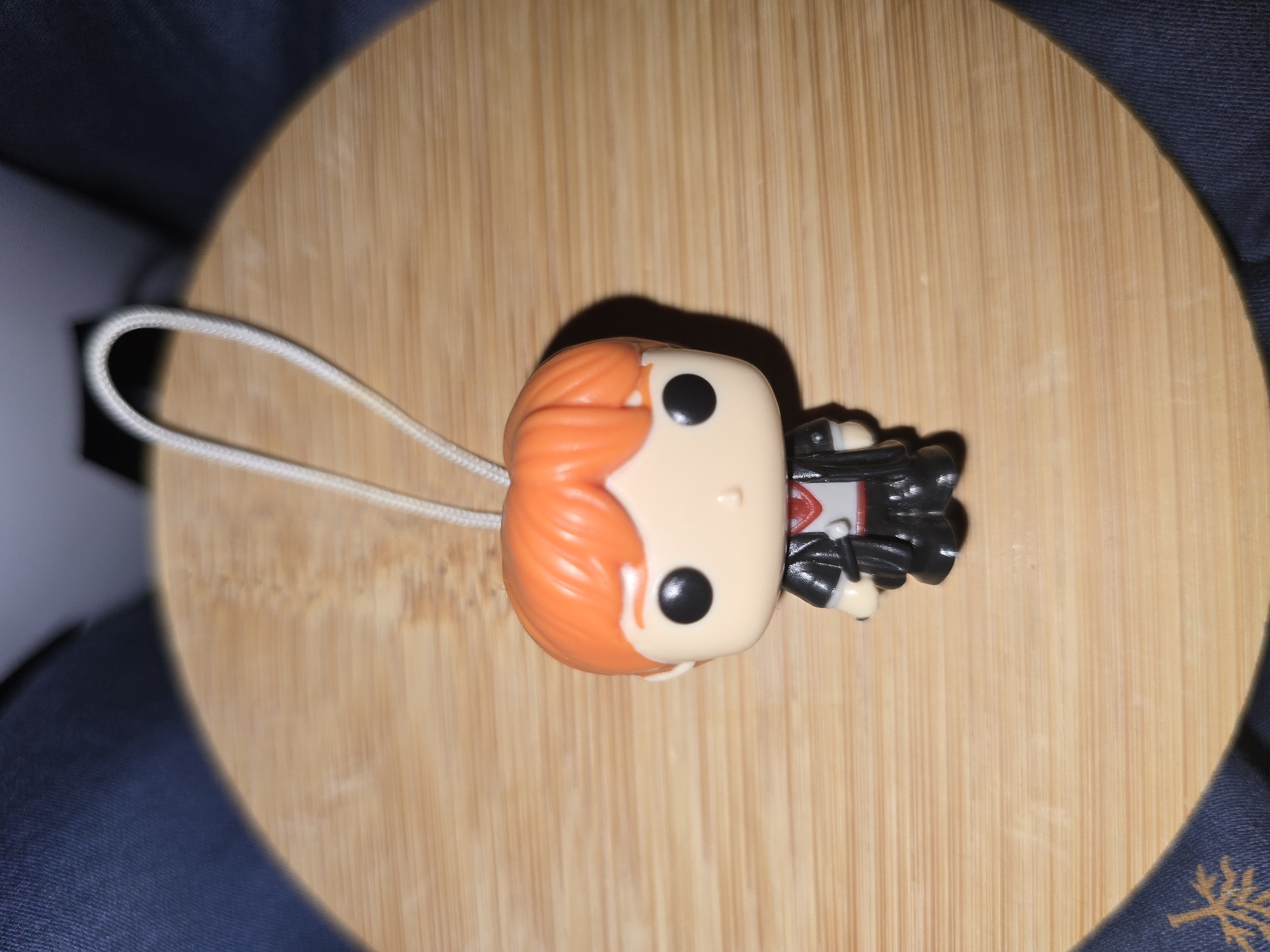 This media file features a close-up, overhead shot of a Funko Pop keychain. The keychain depicts a stylized character with bright orange hair, black eyes, and a black outfit with a white and red detail on the chest. The figure is positioned on a light-colored wooden surface, likely a tabletop or cutting board, with a visible wood grain pattern. To the left of the figure, a white cord, part of the keychain loop, is visible. The background includes a dark blue fabric, possibly clothing or upholstery, with a subtle embroidered pattern of a stylized plant or snowflake in a lighter color. The lighting suggests an indoor setting, with a soft shadow cast by the Funko Pop figure. No specific time of day, weather, or location cues beyond the general indoor setting and the mention of Petropavlivska Borshchahivka, Ukraine, are discernible from the image itself. No text is visible in the media file.