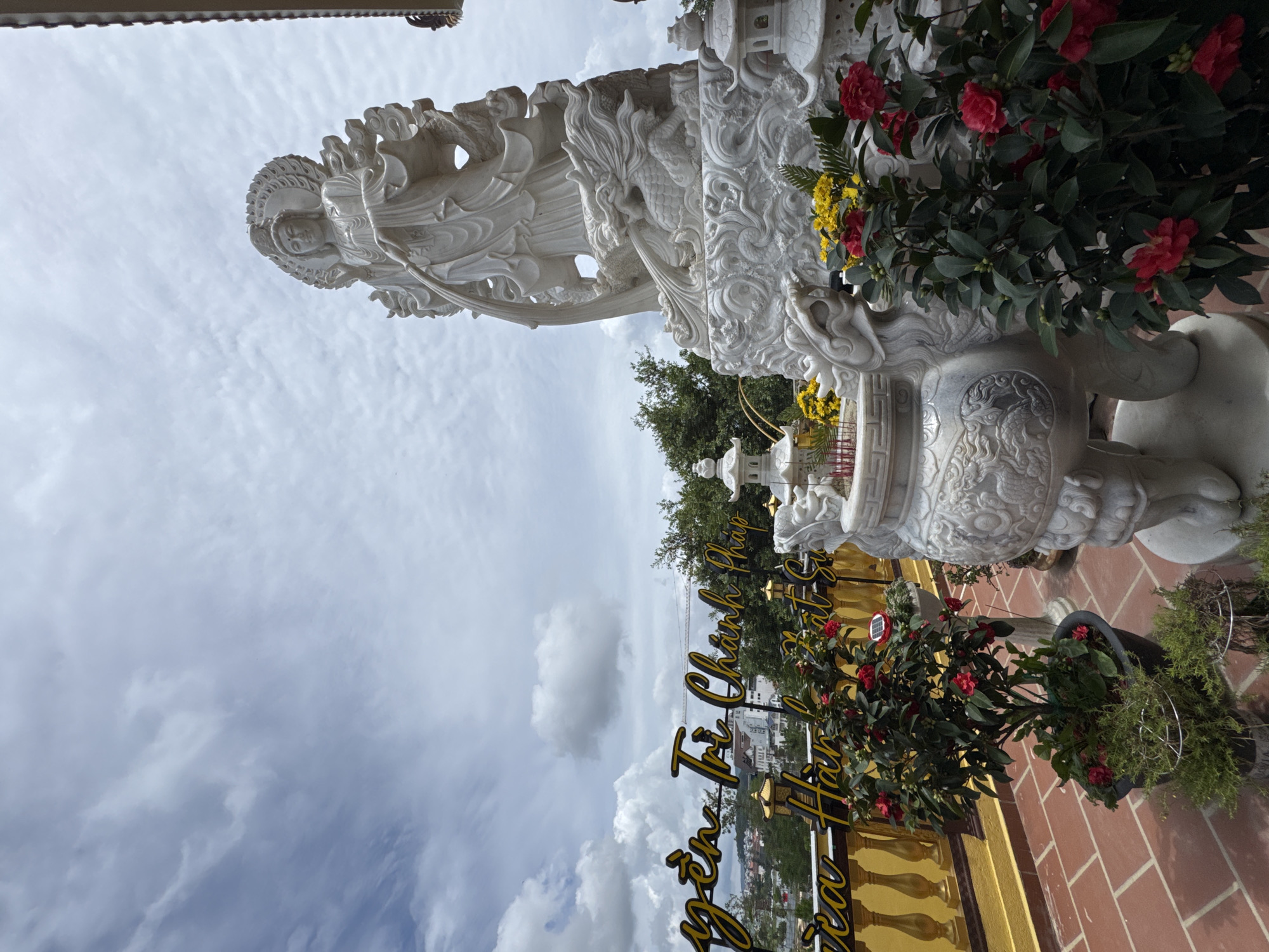 The image depicts a white marble statue of a deity, possibly a Buddhist figure, positioned on an elevated platform overlooking a city. The statue is intricate, with flowing robes and a halo-like headdress, and appears to be supported by a dragon-like creature. In the foreground, there are potted plants with red flowers and decorative incense burners. A yellow railing runs along the edge of the platform. Behind the statue, a sign with yellow lettering on a dark background is partially visible, displaying Vietnamese text. The background shows a cityscape with buildings and lush green trees under a cloudy sky. The location is identified as the Linh Phuoc Pagoda in Da Lat, Vietnam, a popular tourist destination known for its mosaics and architecture. The overall scene conveys a sense of tranquility and cultural significance.