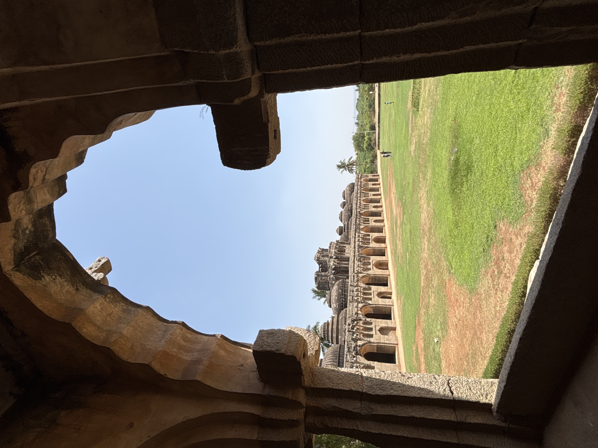 The image captures the Hazara Rama Temple Complex in Kamalapura, India, viewed through a stone archway. A partial view of the sky is visible through the archway's irregular opening. The complex itself is characterized by a long, low structure with arched openings and multiple levels. A grassy field, partially brown, stretches out beyond the structure. Several individuals can be seen on the field in the distance. The foreground consists of the weathered stone of the archway, showing its construction and texture.