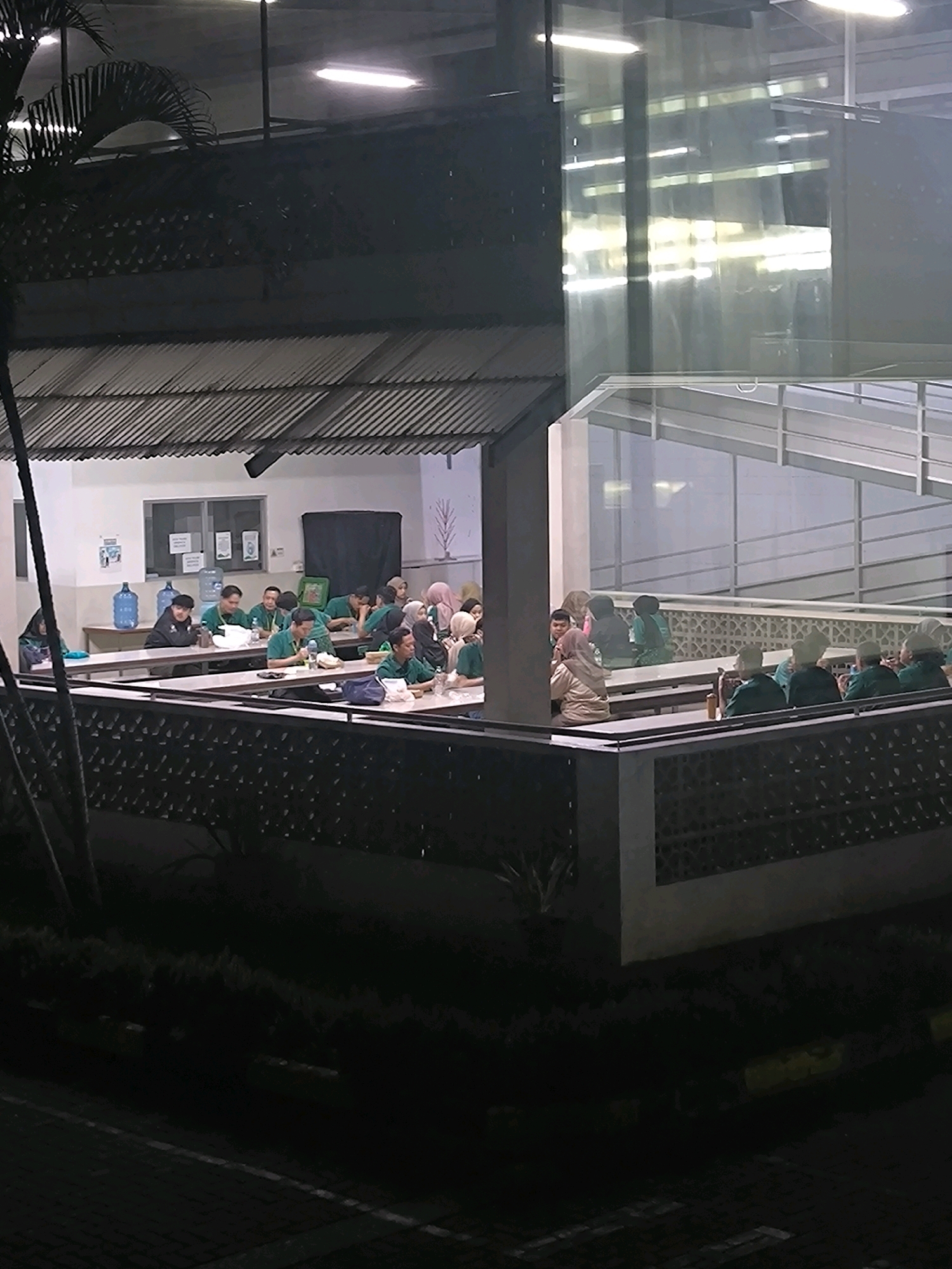 The image depicts an outdoor dining area or cafeteria at night. Several groups of people are seated at long tables, engaged in eating or conversation. Many individuals are wearing green shirts, suggesting a uniform or group affiliation. The setting appears to be part of a larger building complex with a covered roof area over some of the tables and an open, elevated walkway with glass railings in the background. Fluorescent lights illuminate the scene. In the foreground, there are dark bushes and paved ground, suggesting an outdoor or semi-outdoor environment. The architecture features elements like a corrugated metal roof, concrete pillars, and decorative patterned screens. There are also large water dispenser units visible. The overall impression is one of a communal meal or gathering within an institutional or educational facility. The location is not identifiable from the image alone.