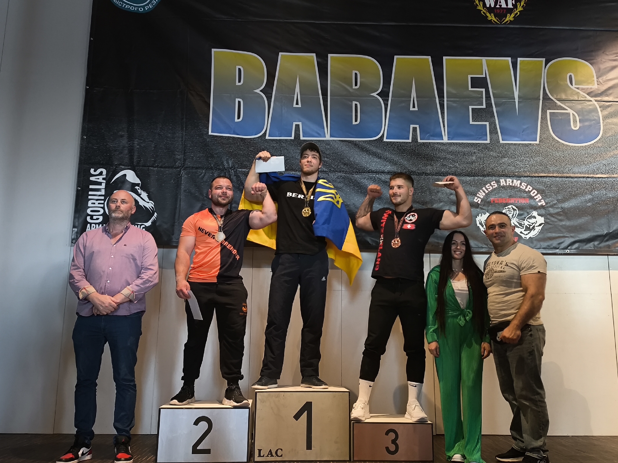 In the city of Romanshorn, Switzerland, a podium ceremony for an arm-wrestling competition is depicted. Three male athletes are positioned on raised platforms labeled '2', '1', and '3' from left to right. The athlete on the first platform (number 2) wears an orange and black t-shirt and black pants. The athlete on the central platform (number 1) is adorned with gold medals and a Ukrainian flag draped over his shoulders, wearing a black t-shirt and black pants. The athlete on the third platform (number 3) is dressed in a black t-shirt and black pants.
Two individuals stand to the left of the podium, a man in a light pink collared shirt and blue jeans, and a man in a beige t-shirt and gray jeans. A woman in a green jumpsuit stands between the third platform and the man in the beige t-shirt. Behind the individuals and the podium, a large black banner with the text "BABAEVS" in large blue and yellow letters is prominently displayed. The environment appears to be indoors, with a plain white wall visible in the background.