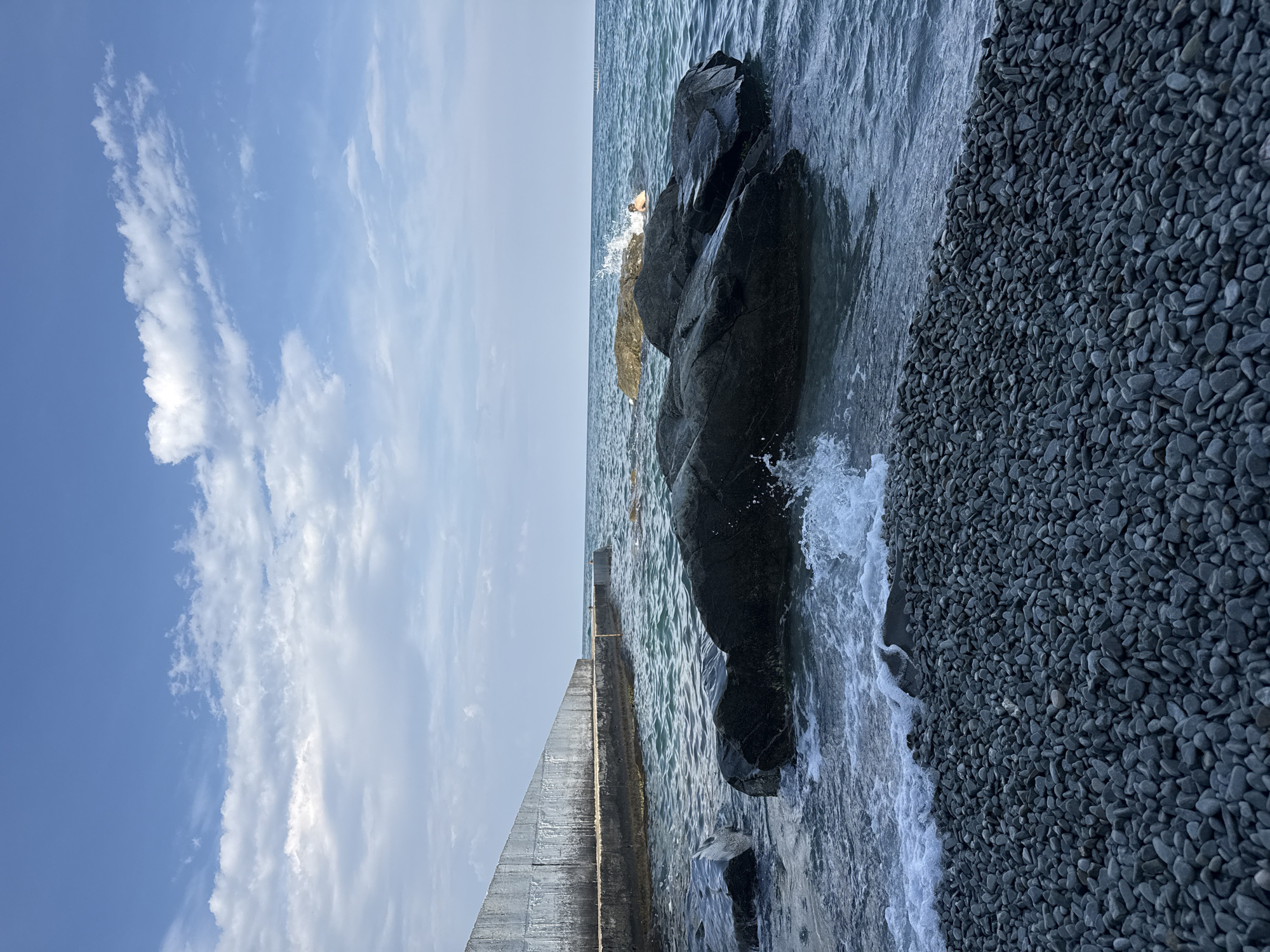 This image depicts a rocky beach in Partenit, Ukraine. In the foreground, a dense expanse of dark grey pebbles lines the shore. The sea in the middle ground is a deep blue, with waves gently lapping against the shore and breaking around large, dark rocks. A concrete structure, appearing to be a seawall or pier, extends from the left side of the frame into the water. In the background, the horizon is met by a clear blue sky dotted with white, fluffy clouds. A single person is visible in the water, partially submerged and appearing to swim or float near a distant rock. The overall scene conveys a peaceful, natural coastal environment.