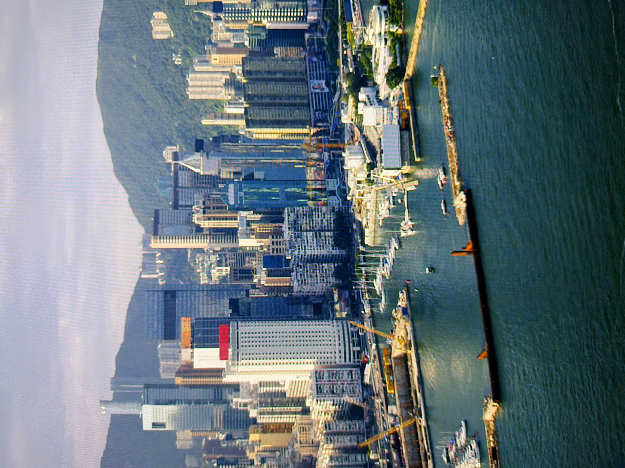 This is an aerial photograph of a harbor and a city skyline, likely in Asia, not Kyiv, Ukraine. The scene features numerous skyscrapers, including a prominent tall white building with a red top. In the foreground, a body of water contains several sailboats and a construction area with cranes and floating structures. A long, dark, rectangular object, possibly a barge or a section of a breakwater, is partially submerged in the water. The background shows a dense cityscape, with mountains visible further in the distance. The sky is overcast. There are no discernible people or animals in the image.