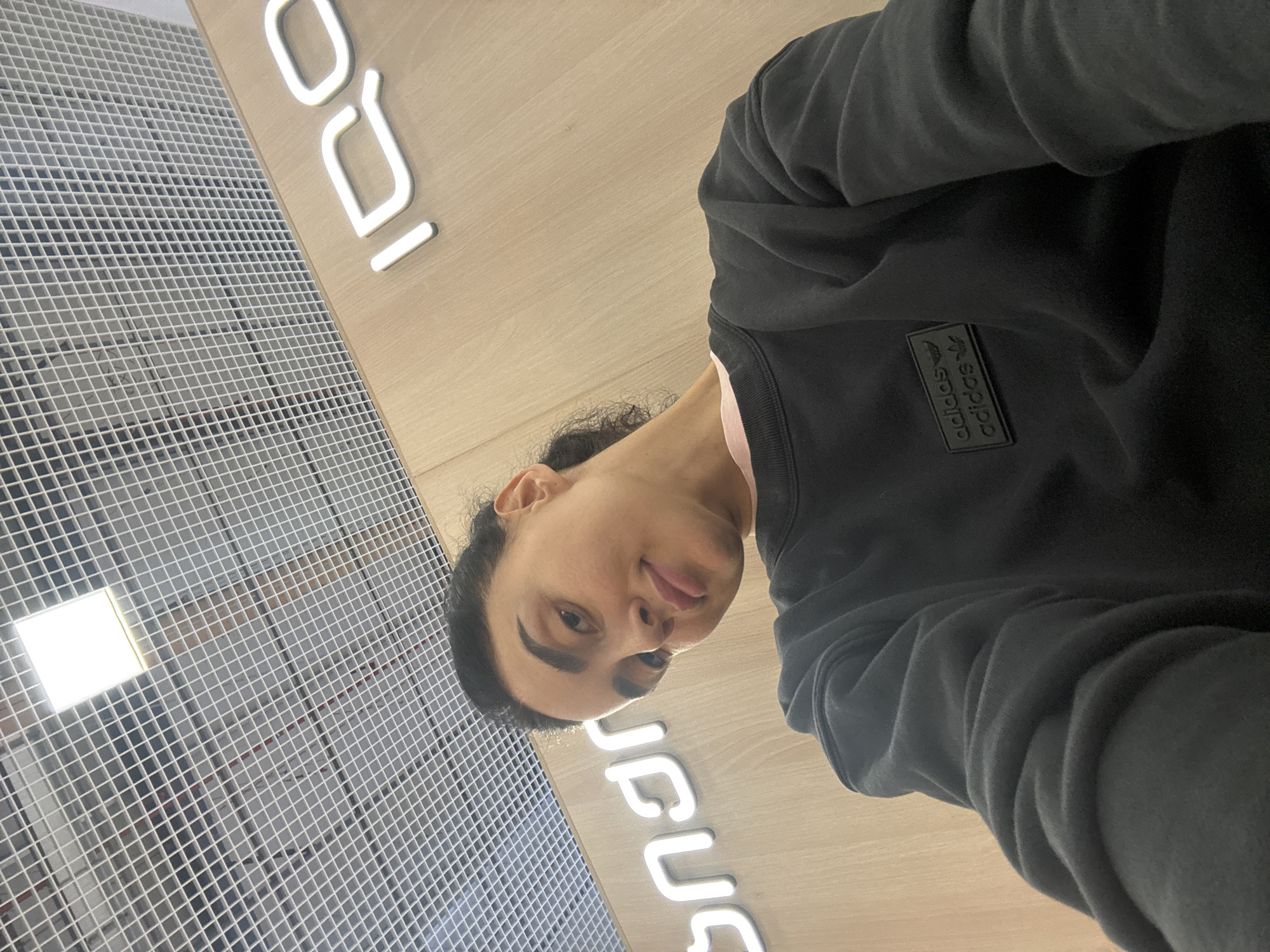 The image is a vertically oriented, close-up selfie or portrait shot inside a modern building, rotated 90 degrees counter-clockwise.
A person with dark hair and a neutral to slightly smirking expression looks directly at the camera. They are wearing a dark, possibly black, long-sleeved sweatshirt. On the upper left chest of the sweatshirt, a black rectangular patch with the "adidas" logo is visible, stacked twice vertically in white text.
The background consists of two distinct sections. To the left of the person's head and body is a light grey grid-like structure, possibly a ceiling or wall panel, with a bright, square light source embedded in it. To the right, behind the person, is a light wooden-textured panel featuring glowing white, three-dimensional letters. These letters appear as "IDQ" (or IQO) in the upper section and "JUUS" (or JCOS) in the lower section, when viewed in the image's current rotated orientation. If the image were rotated clockwise to a standard upright position, these letters would clearly spell "IQOS".
The setting appears to be an indoor commercial space, perhaps a retail store or a public area within a modern building, potentially in Tbilisi, Georgia, given the location context provided. The lighting is bright and even, indicating an interior environment.