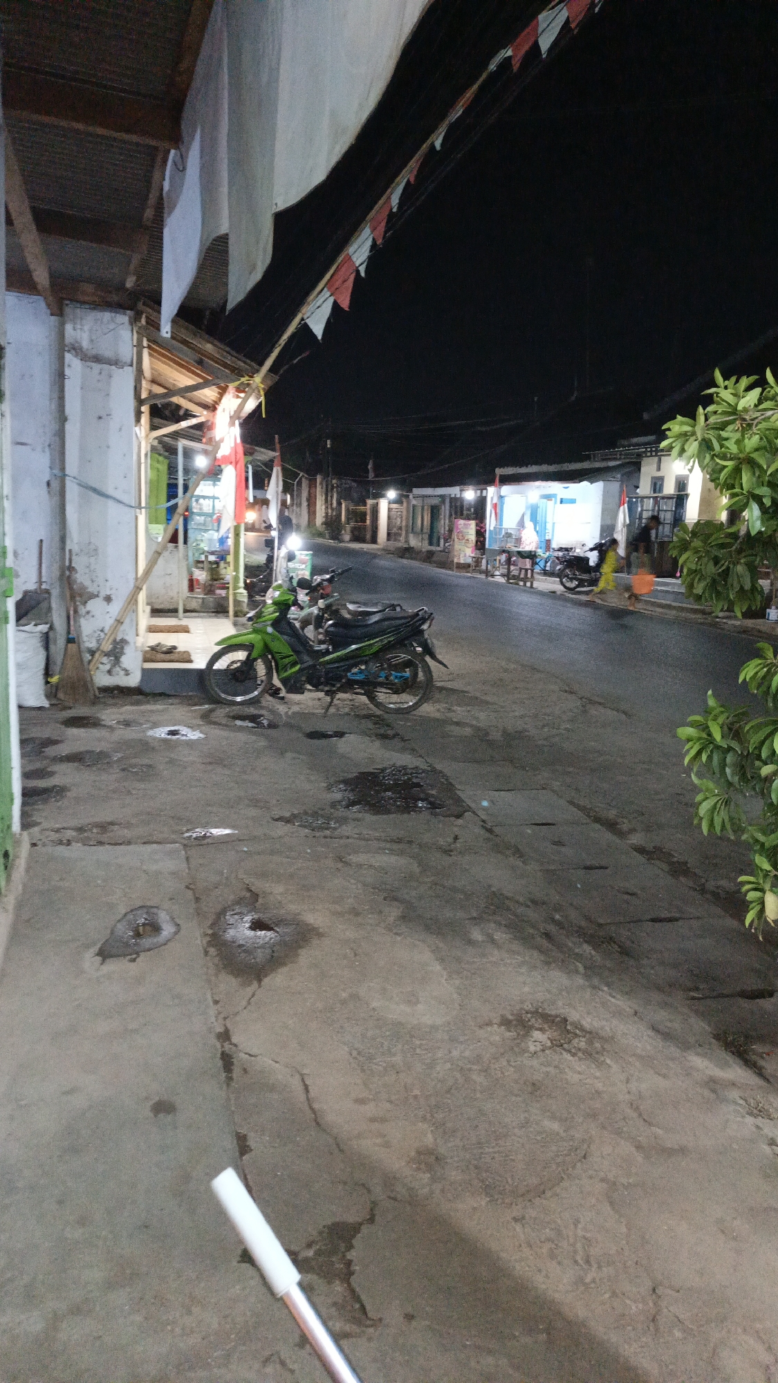 An eye-level, medium shot captures a street scene at night in Bayeman, Indonesia. A green motorcycle is parked on the side of a paved road, with a building to its left and more buildings and a street stretching into the distance on the right. The building on the left appears to be a shop with open storefronts, where flags are displayed. Red and white triangular flags are strung across the street from the left side, above the motorcycle and extending into the dark sky. The ground is concrete, with several damp patches suggesting recent rain or cleaning. A broom and a white bag are leaning against the building on the left. In the distance, several people can be seen walking along the street, and other motorcycles are parked or in motion. Lights from the buildings illuminate the scene. A white object, possibly the handle of a cleaning tool, is in the foreground, partially obscuring the view of the ground. The overall atmosphere is one of a typical Indonesian neighborhood street at night.