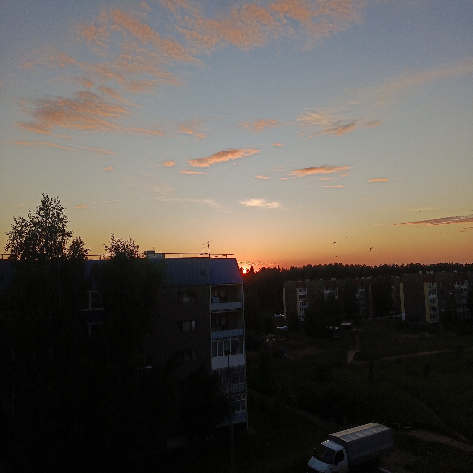 This image depicts a sunset over a residential area in the city of Марковский, Russia. The scene is characterized by a high-contrast lighting, with a brightly illuminated sky transitioning to deeply shadowed foreground elements.
The sky, occupying approximately the upper half of the frame, displays a gradient of colors. The upper sky is a light, clear blue, transitioning downwards into vibrant hues of orange, amber, and deep red near the horizon. Scattered, elongated, and irregular altocumulus clouds, illuminated by the setting sun, appear pinkish-orange against the blue and transitional sky. The sun itself is visible as an intense orange-red disc, partially obscured by a distant tree line and a building on the horizon. A faint, linear contrail is visible in the upper-left quadrant of the sky. Multiple small, dark avian forms are discernible in flight against the bright sky, primarily in the central-right portion of the frame.
The midground features a prominent multi-story residential building on the left, possessing a distinct blue, slightly gabled roof. The building's upper facade, including a roof railing and antenna-like structures, is silhouetted against the bright sky. The building itself is largely in shadow, though windows and balconies are discernible. To the right, behind a dense tree line, several other multi-story residential buildings of uniform block-style architecture are visible. These buildings are also predominantly in deep shadow, with visible windows and balconies.
The foreground and lower midground are characterized by deep shadows, rendering most details indistinct. Silhouettes of numerous trees and dense bushes dominate this area. On the far lower right, a light-colored delivery or cargo van, possibly white or light grey, with a white canvas or tarpaulin cover over its cargo bed, is partially visible, parked on an unlit surface. No human figures are discernible in the scene.
