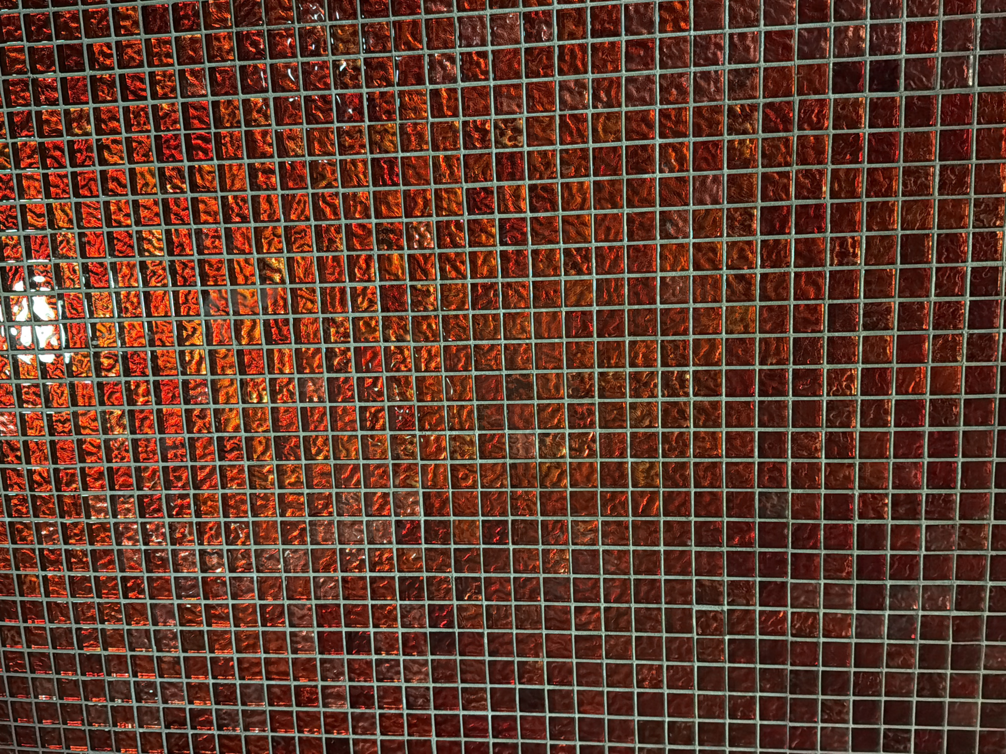 The image shows a close-up of a wall covered in small, square, red mosaic tiles. The tiles have a slightly irregular surface texture, which causes light to reflect off them in various ways, creating a shimmering effect. The grout between the tiles appears to be a light gray or silver color. The lighting in the image is warm and appears to be coming from above, highlighting the red and orange tones of the tiles. There are no people or identifiable objects in the image, and it is a still shot of a textured surface. The location in Thuận Hóa Ward, Vietnam is not discernible from the image itself.