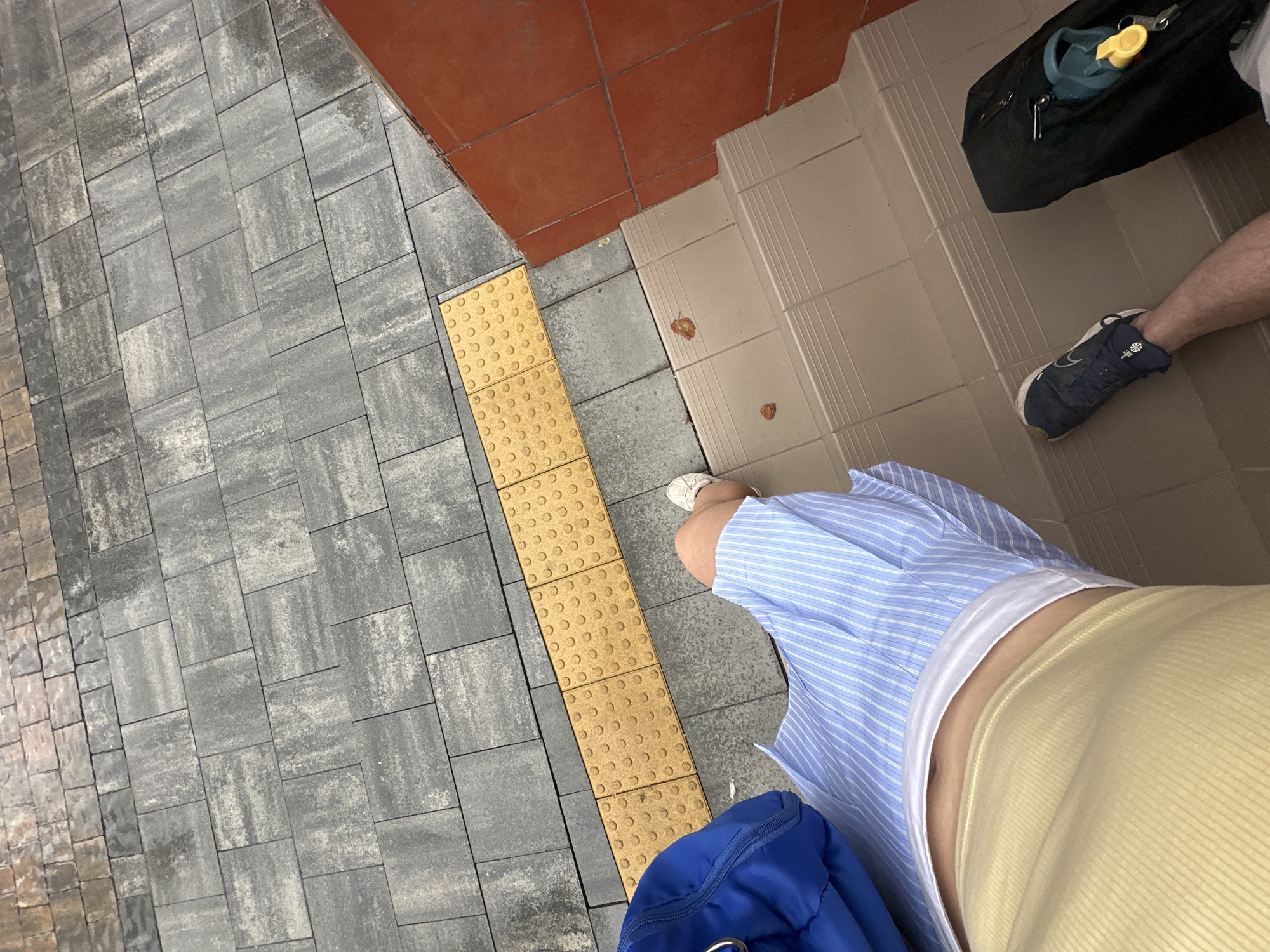 The image shows a first-person perspective of a person standing on stairs. The person is wearing a yellow top, a blue and white striped skirt, and blue sneakers. To the left is a blue duffel bag. To the right is a black suitcase with a green object and keys attached. The stairs are made of light-colored tiles. Above the stairs, the ground is paved with grey and brown rectangular tiles, with a yellow tactile paving strip in between. There are two dry leaves on the stairs. The context of Ternopil, Ukraine, is not directly evident from the visual information in the image.