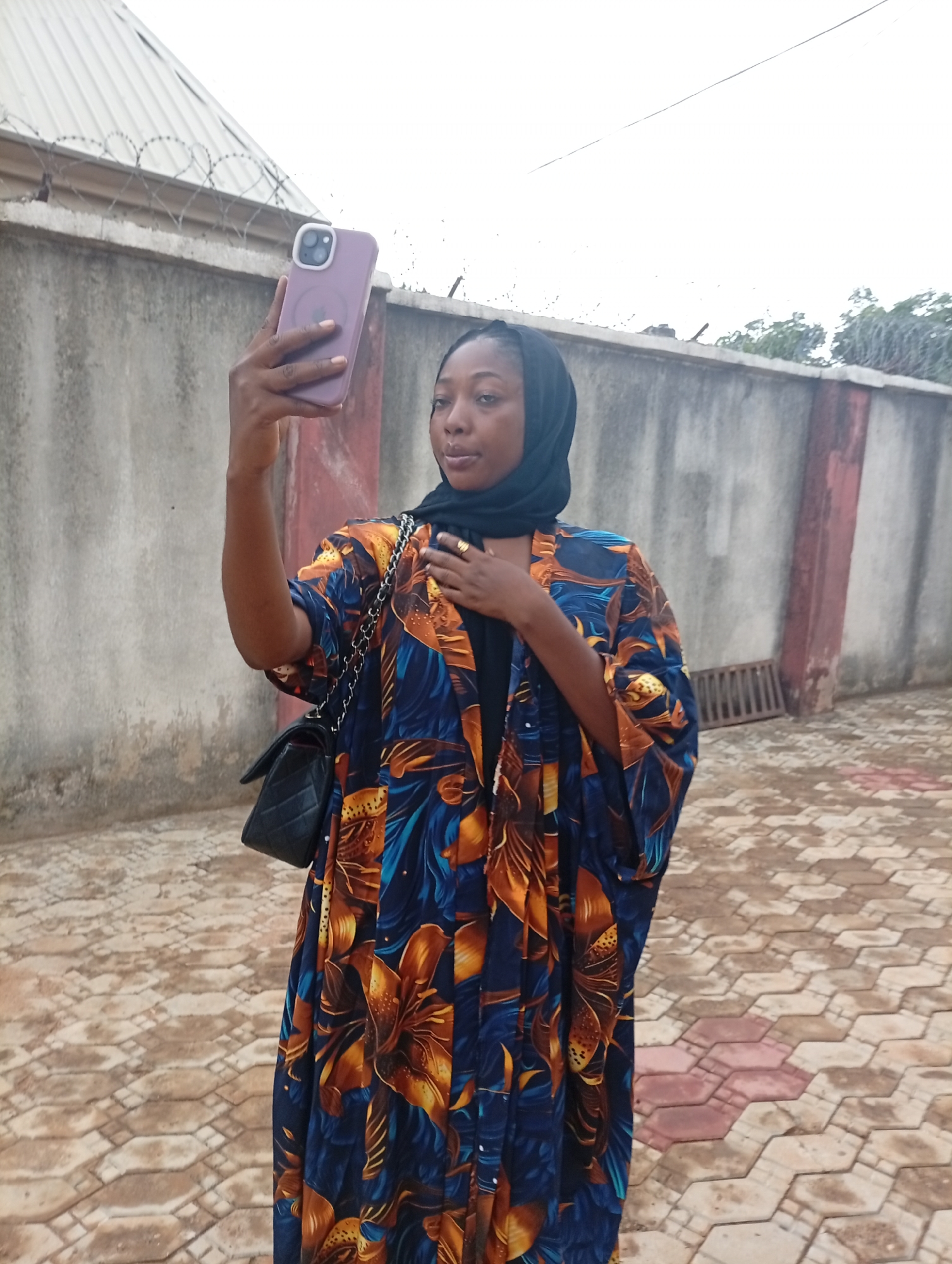 A woman stands on a paved surface taking a selfie with a lavender-colored smartphone. She is wearing a dark headscarf, a blue and orange floral patterned robe, and a black purse hangs from her shoulder. She is holding her hand to her chest. Behind her is a concrete wall topped with barbed wire and a small building. The location is in the city of Unknown, Unknown.