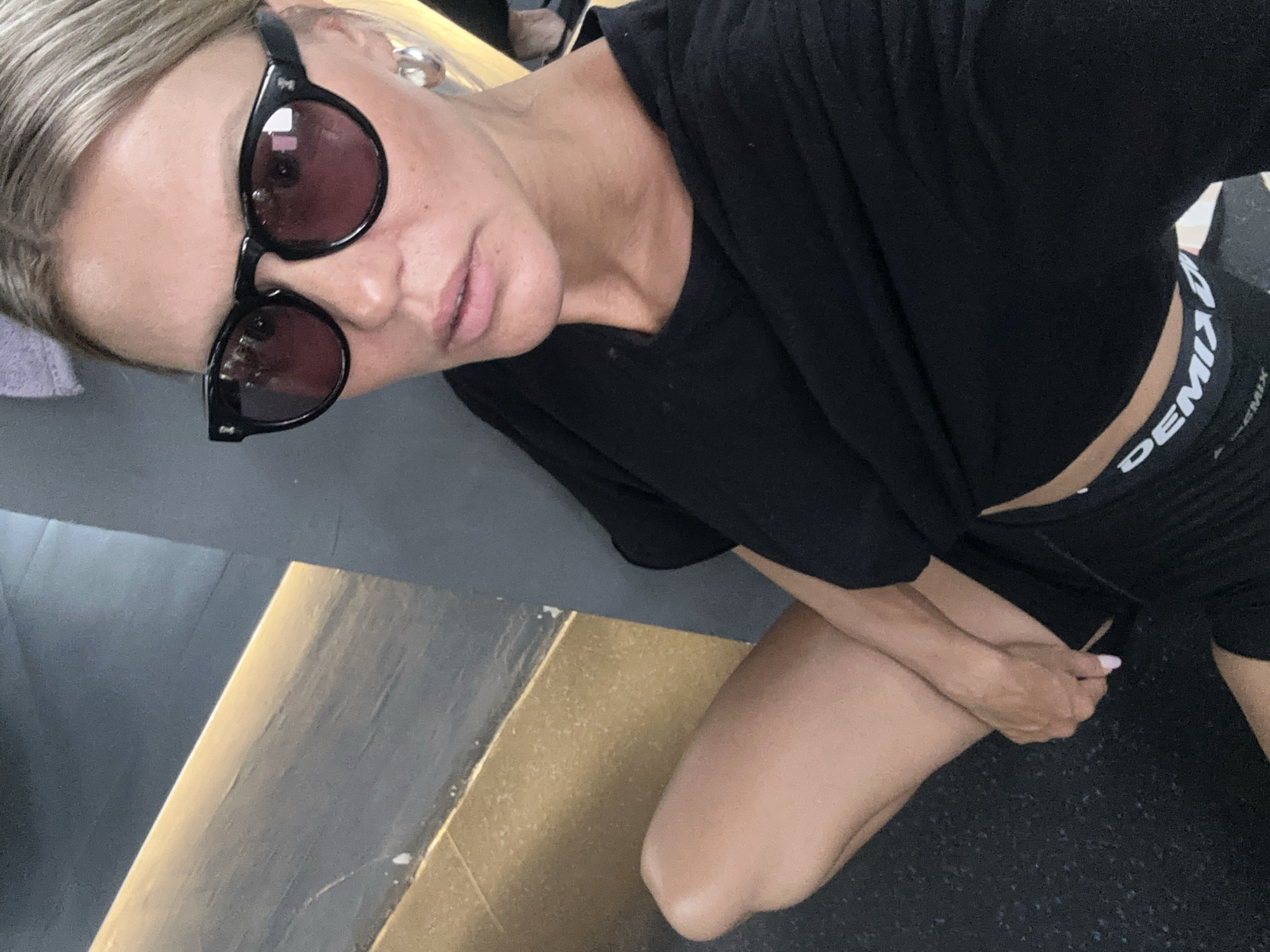 The media file shows a woman wearing black athletic shorts and a black t-shirt, with a black branded waistband visible. She is wearing dark sunglasses. Her hair is blonde and styled back. The woman is sitting on what appears to be a gym floor, which is textured and dark gray. To her left, there is a raised surface with a golden, metallic finish, possibly part of gym equipment or the gym's interior design. The lighting suggests an indoor environment, likely during the daytime. The woman's posture and attire suggest she might be resting during or after a workout. The visible text on the waistband reads "DEMIX".