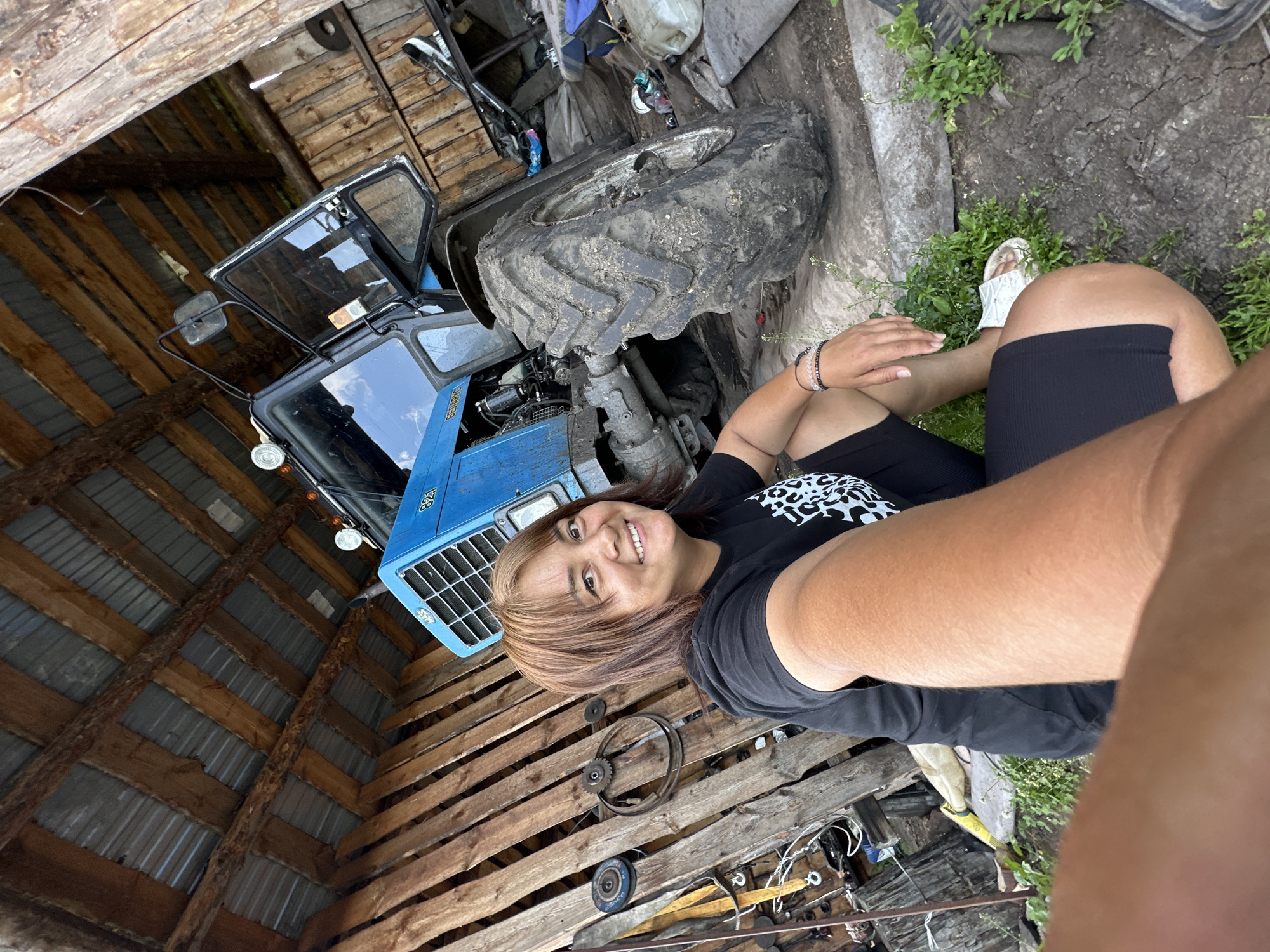 A woman with shoulder-length brown hair, wearing a black t-shirt with a white leopard print design and black shorts, poses for a selfie in an outdoor, rustic setting. She is smiling broadly and looking directly at the camera. Her left arm is extended towards the camera, partially obscuring the view. She is wearing white slides on her feet and several bracelets on her left wrist.
The setting appears to be a farm or workshop, with a blue tractor prominently featured. The tractor is partially covered by a large, muddy tire. The background includes a wooden structure with a corrugated metal roof, suggesting a barn or shed. Wooden pallets are stacked on the ground. Various tools and mechanical parts are scattered around the scene, including gears and a belt. Green foliage is visible in the upper right portion of the image, indicating an outdoor environment. The lighting suggests it is daytime, possibly overcast, as there are no strong shadows. The overall mood is casual and relaxed, with the woman appearing happy and comfortable.
