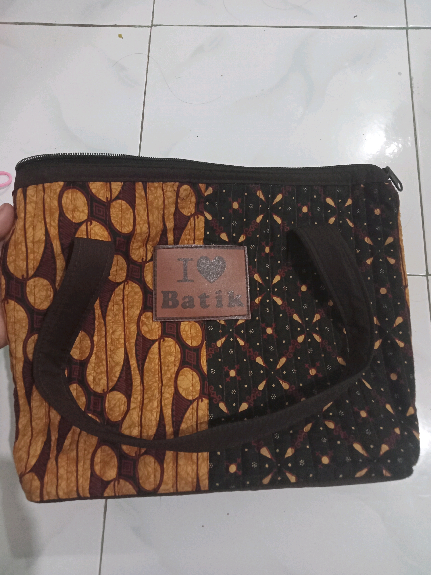 A close-up, overhead shot shows a brown batik fabric bag lying on a tiled surface. The bag has a zipper closure along the top and two brown fabric handles. A small, square patch is sewn onto the front of the bag, bearing a heart symbol above the word "Batik" in capital letters. The left half of the bag features a pattern of large, abstract shapes in yellow and dark red against a lighter brown background, while the right half has a repeating pattern of small, intricate motifs in yellow and dark red on a black background. A person's hand with a pink accessory is partially visible on the left side of the image, holding the bag. The tiled floor appears to be white with faint grey grout lines, suggesting an indoor setting, possibly in Sukomananti, Indonesia.