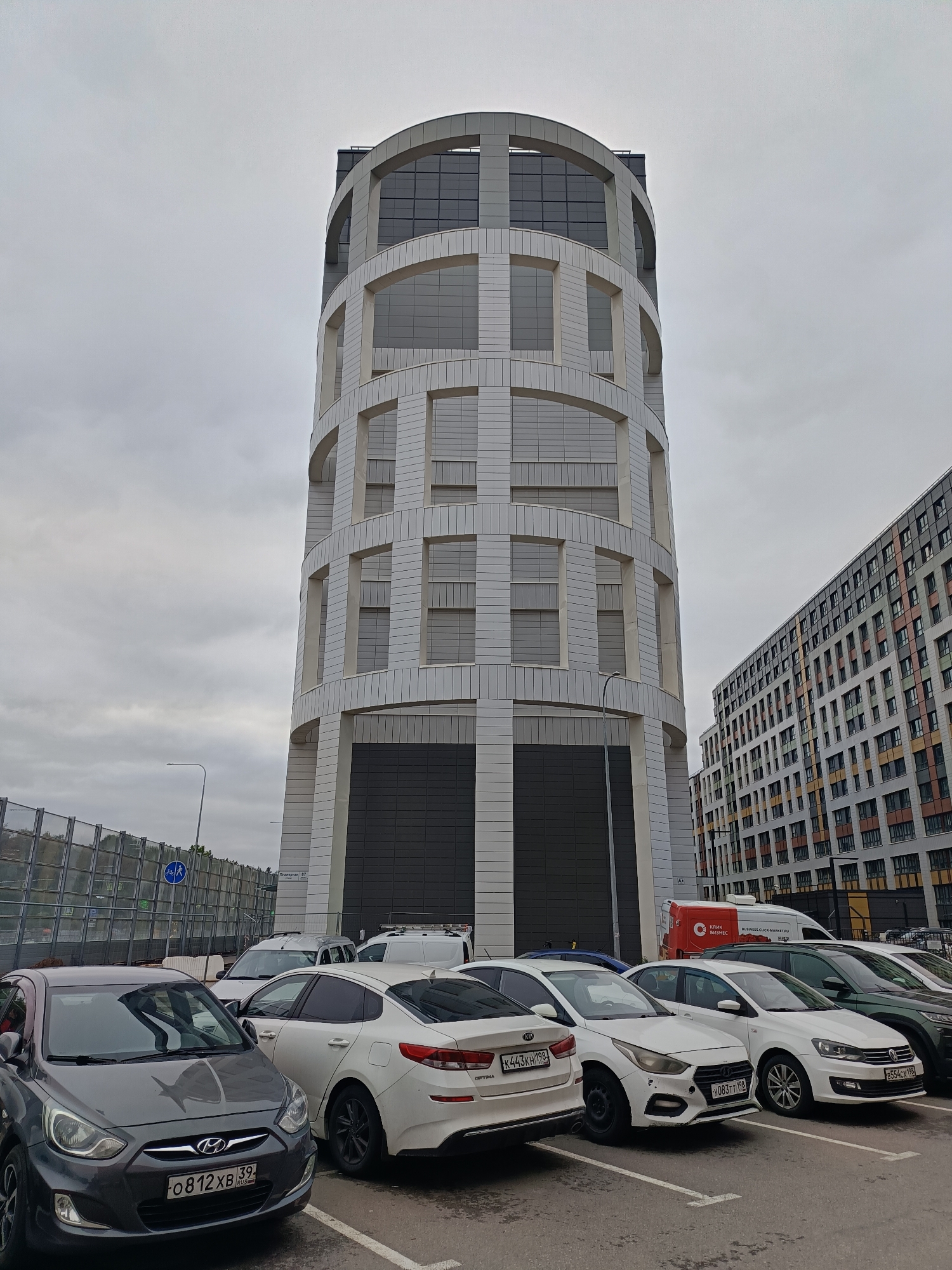 The image depicts a cylindrical building with a distinctive architectural design, featuring a grid-like pattern of white tiles and arched windows. It is situated in Saint Petersburg, Russia, as indicated by the Russian license plates on the cars parked in front. The building is tall, stretching towards the overcast sky, and has a dark base. In the foreground, several cars are parked in a designated parking area. A multi-story apartment building is visible on the right side of the frame, and a noise barrier lines the left side, suggesting a nearby roadway. A red van with a logo is parked behind the vehicles. The overall scene appears to be an urban setting on a cloudy day.