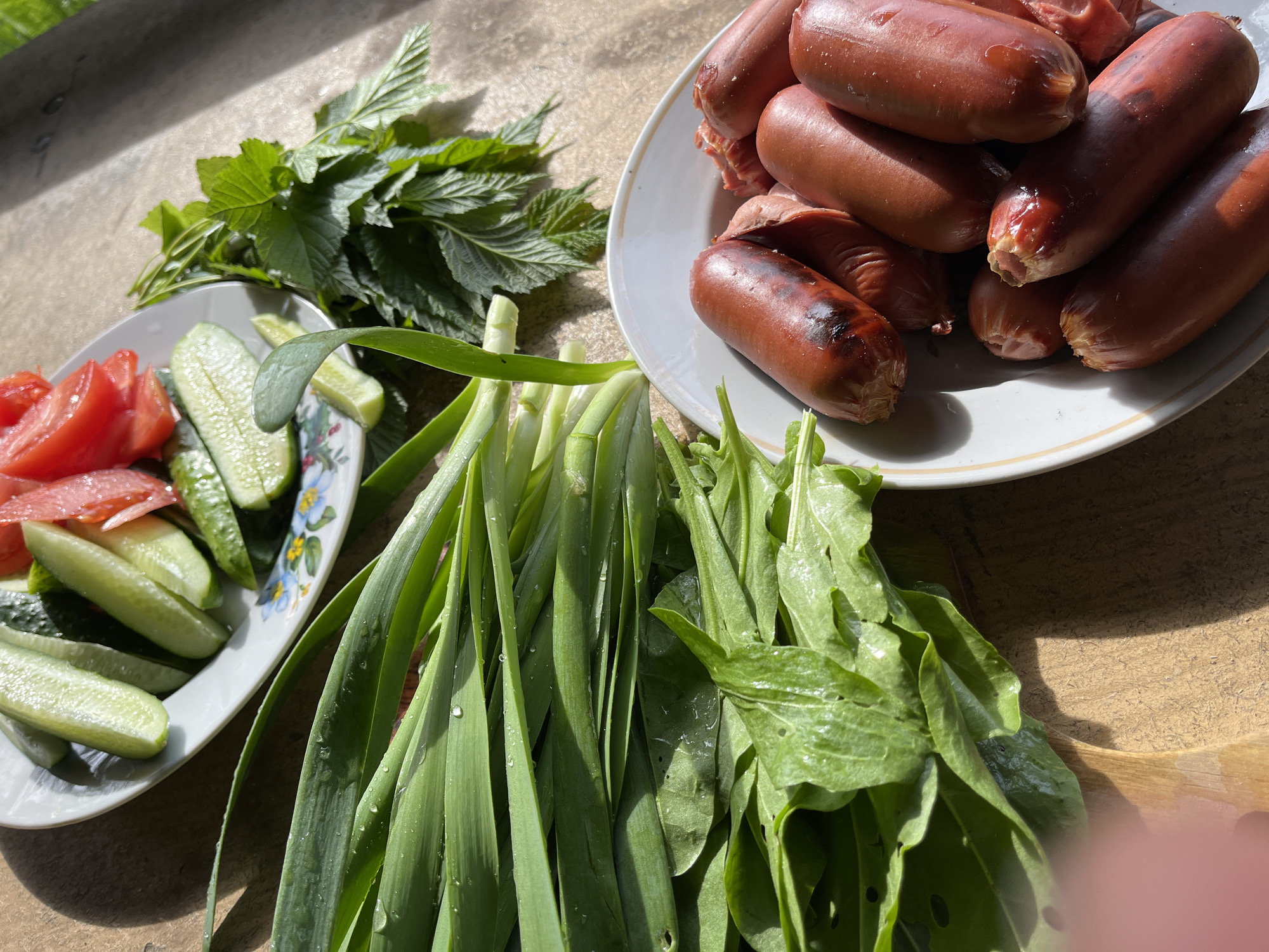On a sunlit wooden surface in Chaikovsky, Russia, lies a meal arrangement. A white plate holds a pile of browned sausages. Adjacent is another white plate containing sliced tomatoes and cucumbers. Bunches of green onions, sorrel and herbs are positioned beside the plates.