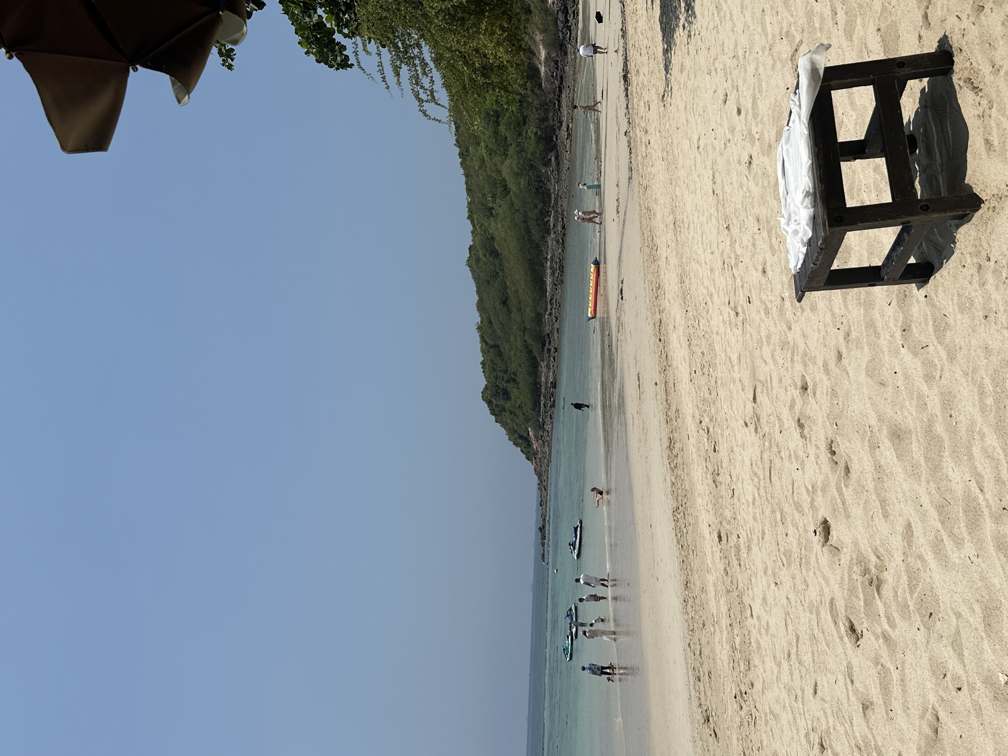 The image captures a sunny beach scene in Pattaya City, Thailand. The beach is sandy and dotted with footprints. In the water, several people are seen standing near jet skis, while others are walking along the shoreline. In the foreground, there is a small, dark wooden stool with a white cloth draped over it. The sky is clear and blue. The beach is bordered by a green, tree-covered area. A portion of a beach umbrella is visible in the upper left corner of the image.