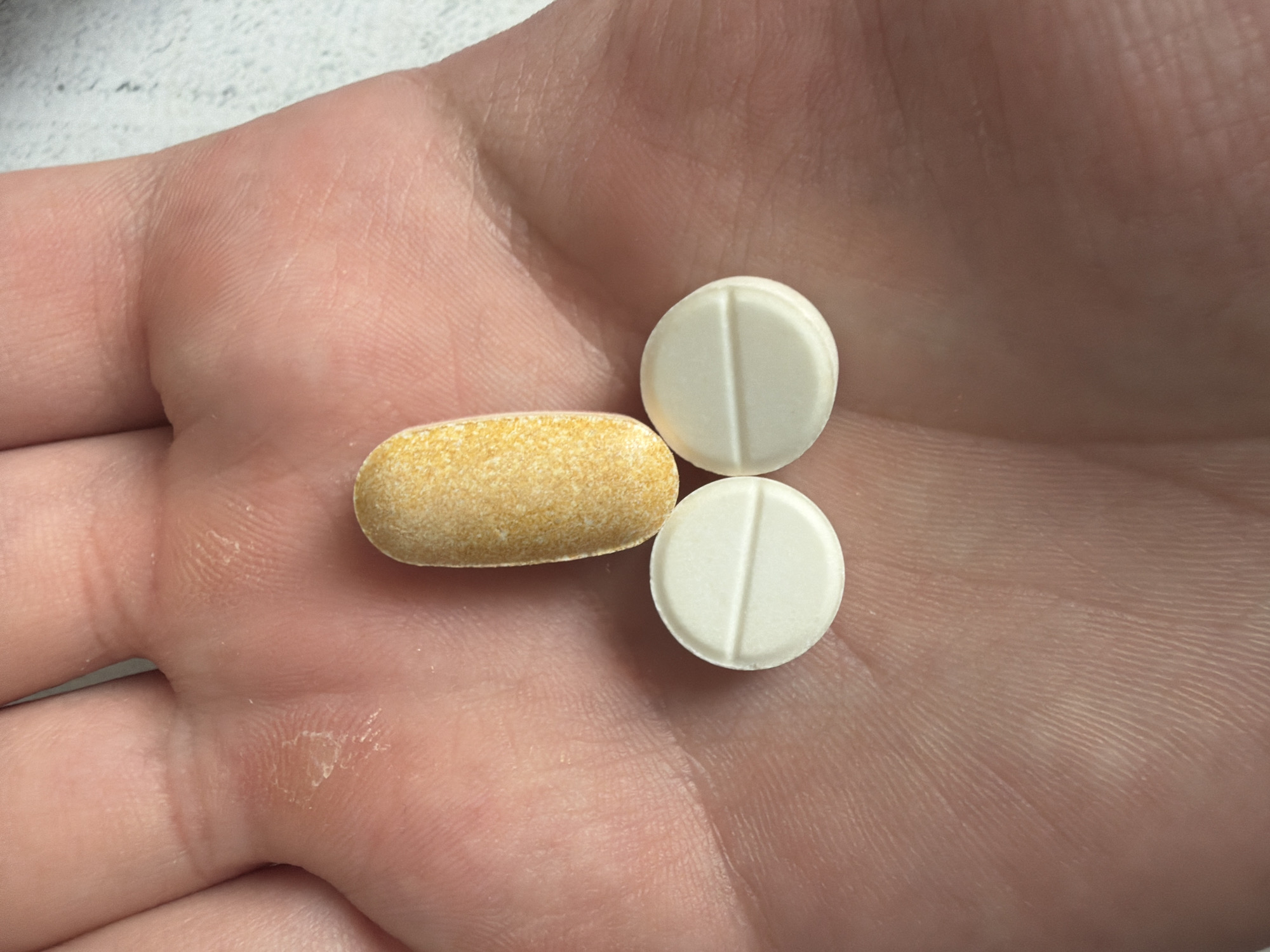 The image shows three pills placed on a human hand. One pill is oval and orange-yellow, while the other two are round and white. The pills are arranged in a phallic shape. The hand appears to be that of an adult and shows visible skin texture and creases. The background is out of focus and appears to be a light-colored, possibly textured surface. There is no indication of the city of Shymkent, Kazakhstan, or any specific location.