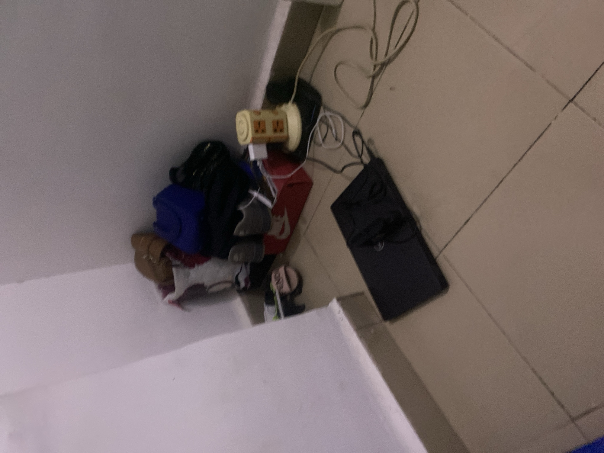 The image depicts a cluttered corner of a room, likely a bedroom or storage area, with items piled on the floor and against the wall. In the far right of the image, a yellow, cylindrical power strip with multiple outlets is plugged into a wall socket. Various chargers and cables are connected to the power strip, some of which are coiled on the floor. To the left of the power strip, a red Nike shoebox is visible, containing what appears to be a pair of gray sneakers. Behind the shoebox, a white plastic bag holds some clothing items, including a blue garment and a brown shoe. Further to the left, a black laptop computer is placed on the tiled floor, open with its screen facing upwards. A pair of black flip-flops with the word "FENDI" visible on them rests near the laptop. The walls are painted a light color, and the floor is covered in beige square tiles. The scene suggests a lived-in space with items that are frequently used or stored. The location is specified as Howayi, Nigeria.