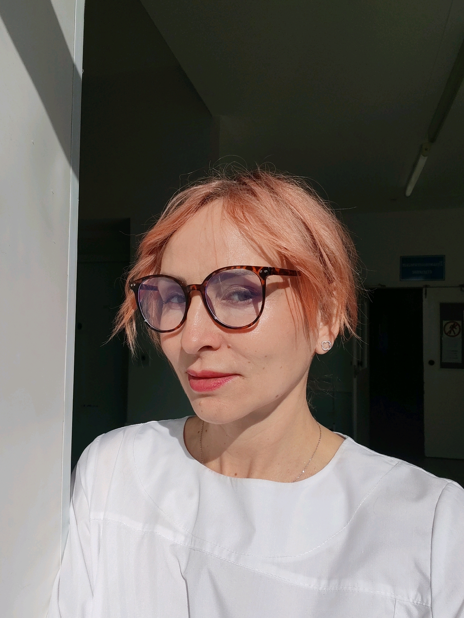 A woman with short, peachy-pink hair and glasses is positioned in the foreground, looking slightly to the left. She is wearing a white garment, possibly a lab coat, with a delicate chain necklace. Her face is illuminated by sunlight coming from the left, casting a shadow on the right side of her face and neck. Behind her, the environment appears to be an indoor setting with doorways and signs visible in the dimly lit background. The overall scene suggests a professional or institutional location, possibly within a hospital or clinic, within the city of Chaikovsky, Russia.
