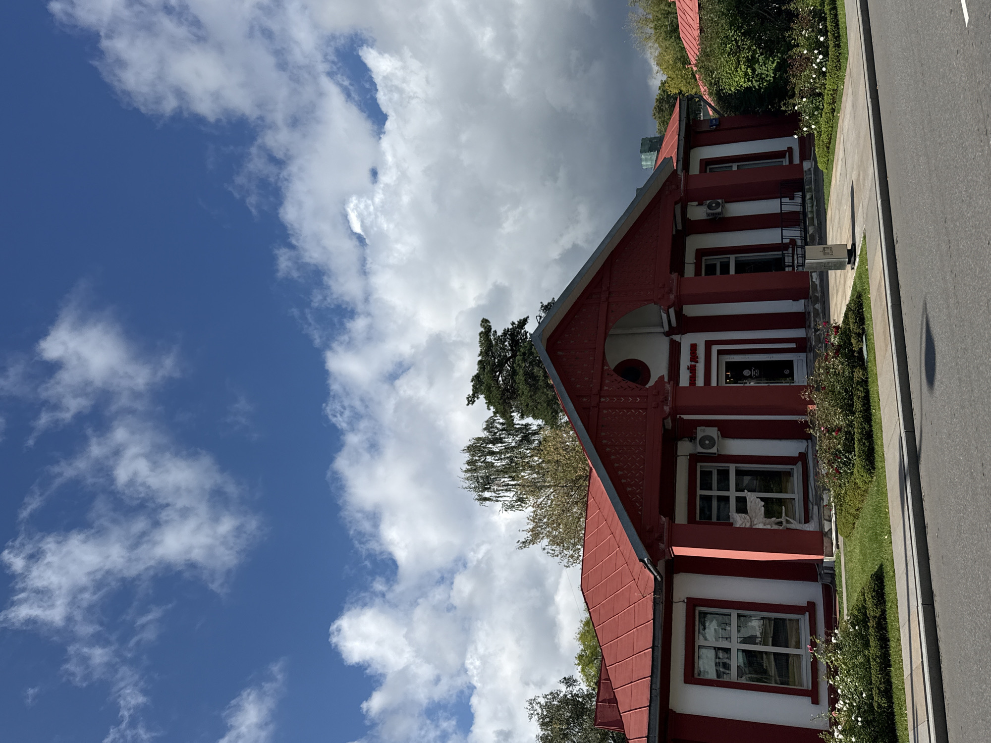 The image shows a red and white building with a red roof, set against a bright blue sky with white clouds. The building has several windows and is flanked by trees and bushes. In front of the building, there is a sidewalk and a street. A trash can is visible on the sidewalk. The building appears to be a restaurant or cafe.
The building is located at Prospekt Mira, 114a, Moscow, Russia. It is a historic building that was once part of the Ostankino Palace estate. The building is now home to a restaurant called "Dom Restaurant". The restaurant is known for its traditional Russian cuisine and its beautiful setting. The building is surrounded by a garden with rose bushes and trees. The street in front of the building is lined with trees and there is a sidewalk for pedestrians. The sky is blue with white clouds, indicating a pleasant day.
