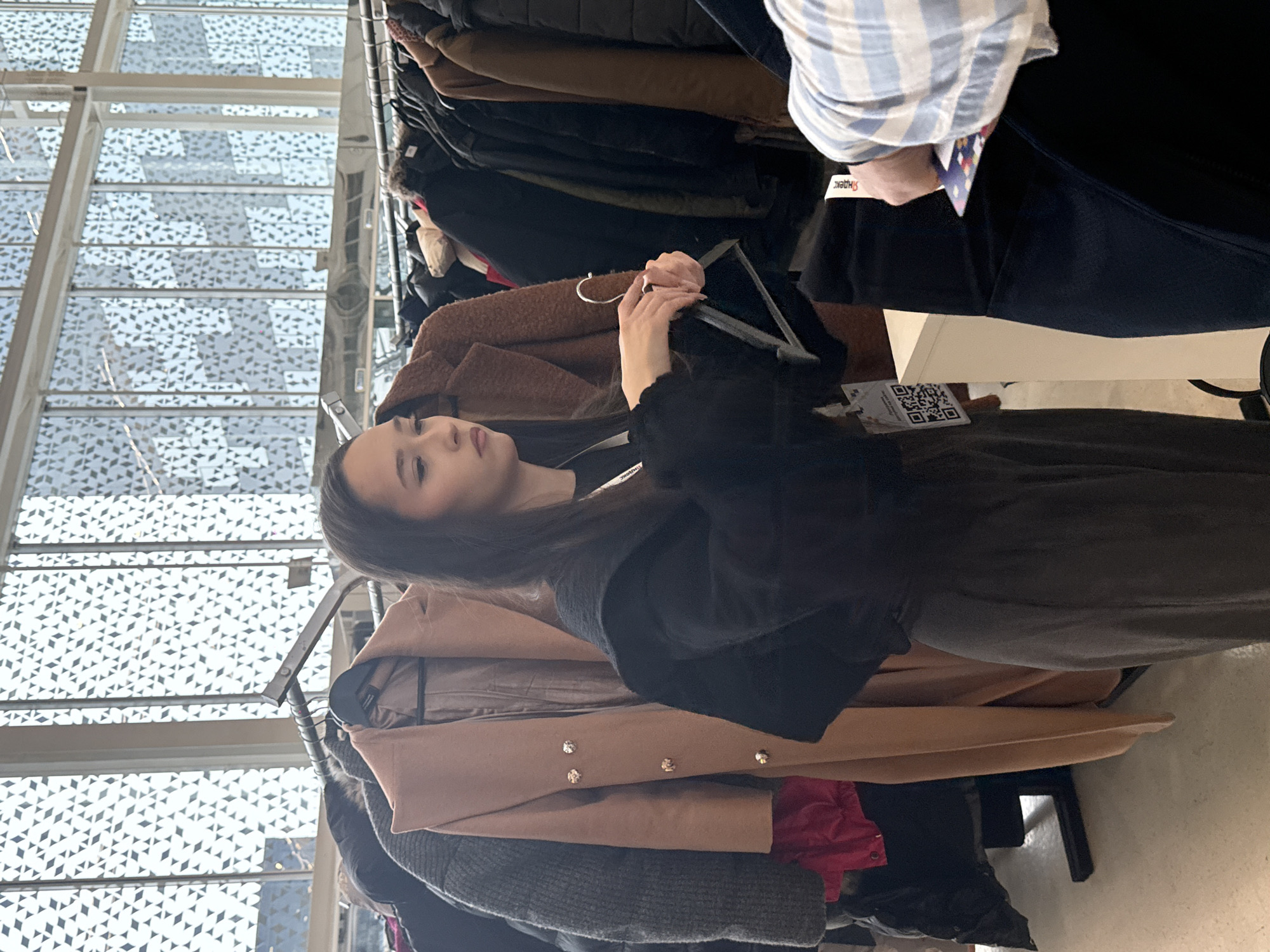 The image, taken in Moscow, Russia, captures an indoor scene of individuals browsing clothes. The primary subject is a young woman with long dark hair, positioned centrally and looking down at a dark garment she holds on a hanger. She wears a dark long-sleeved top or jacket and dark trousers, and her expression is neutral and focused.
To her right, partially visible from the mid-torso down, is another person wearing a light blue and white striped long-sleeved shirt and dark trousers. A white wristband with red text that appears to read "Shabac" is visible on their wrist.
Both individuals are standing near a metal clothes rack filled with various outerwear, including coats and jackets in shades of brown, black, grey, and beige. A prominent light brown coat with two gold-colored buttons is noticeable towards the bottom of the rack. The presence of these clothes racks and multiple garments suggests a retail environment, such as a clothing store, market, or a pop-up event.
In the lower right, on a surface or table, a white card with a black QR code is visible, along with some small, unreadable text beneath it. The background features a striking architectural design: large windows or a glass ceiling composed of multiple panels, each with a repeating geometric pattern of light-colored, possibly translucent, diamond or rhomboid shapes. This bright, natural lighting indicates it is daytime. The overall setting is a modern, spacious interior, characteristic of contemporary urban design.