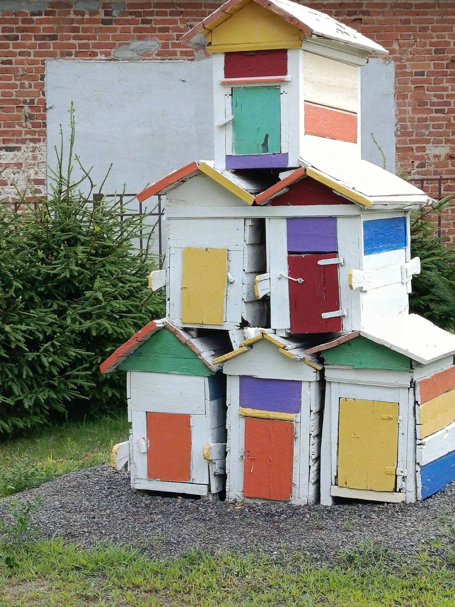 A stack of colorful, weathered wooden beehives stands in front of a rough brick wall and some evergreen trees. The beehives are painted in various bright colors, including red, yellow, blue, green, and purple, with white as the base color. Each hive has a small door, also painted in different colors. The stack is uneven, with some hives leaning more than others. The ground in front of the beehives is covered with gravel and some green grass. In the background, a large section of the brick wall is covered with a lighter colored, textured material, possibly stucco. A portion of a metal fence can be seen behind the evergreen trees. The overall scene suggests an outdoor apiary or a decorative display of beehives.