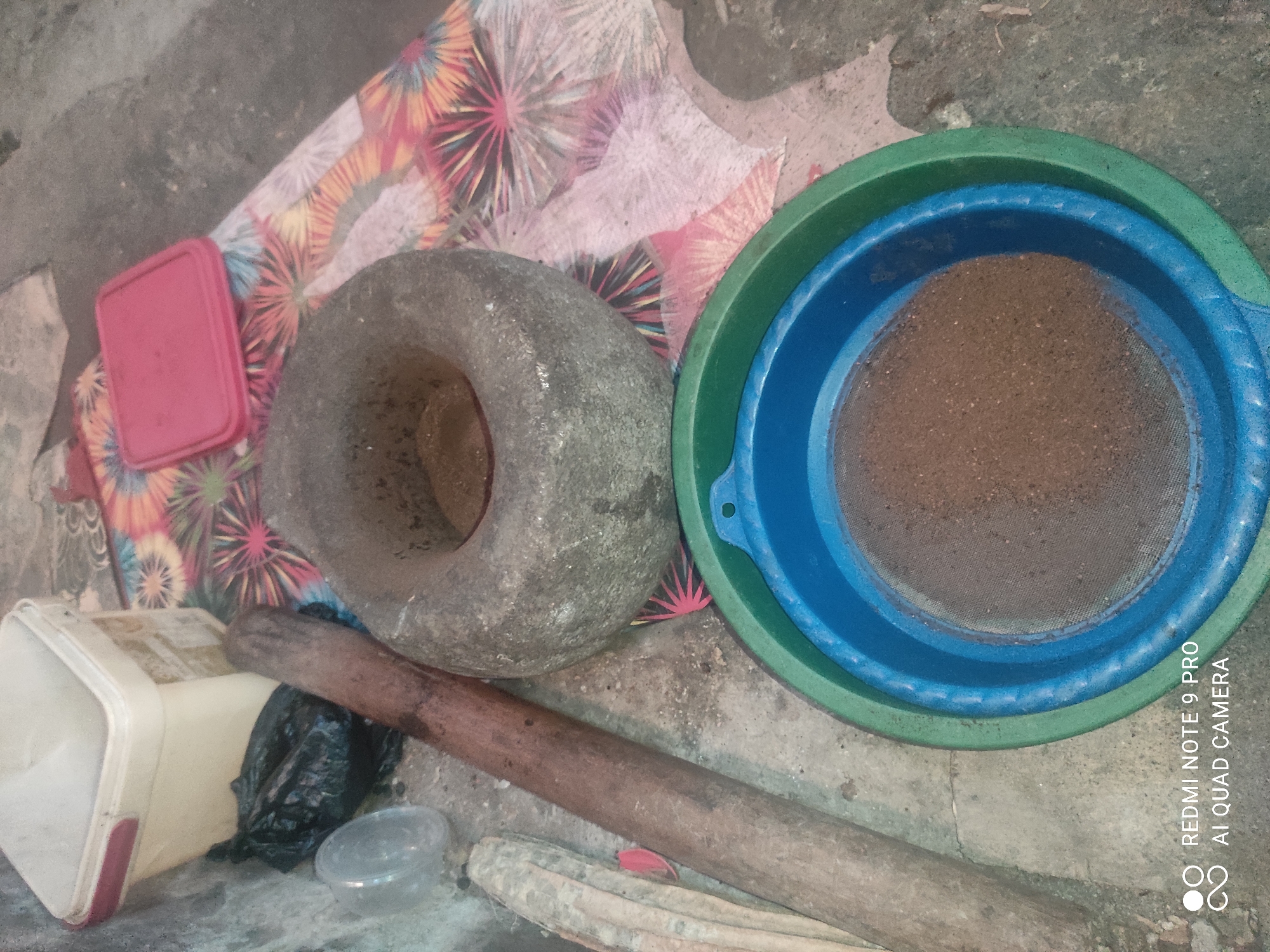 A mortar and pestle, along with a sieve and a plastic container, are arranged on the ground in Sakra, Indonesia. The mortar is a large, rough stone with a concave interior, and the pestle is a long, wooden object. A blue sieve, filled with a brown granular substance, is nested inside a larger green plastic bowl. To the left of the mortar, a white plastic tub with a pink lid sits next to a black plastic bag and a clear plastic lid. A colorful, patterned cloth is visible beneath some of the objects. The ground appears to be a rough, concrete surface. The lighting suggests an outdoor environment, possibly a yard or alleyway.